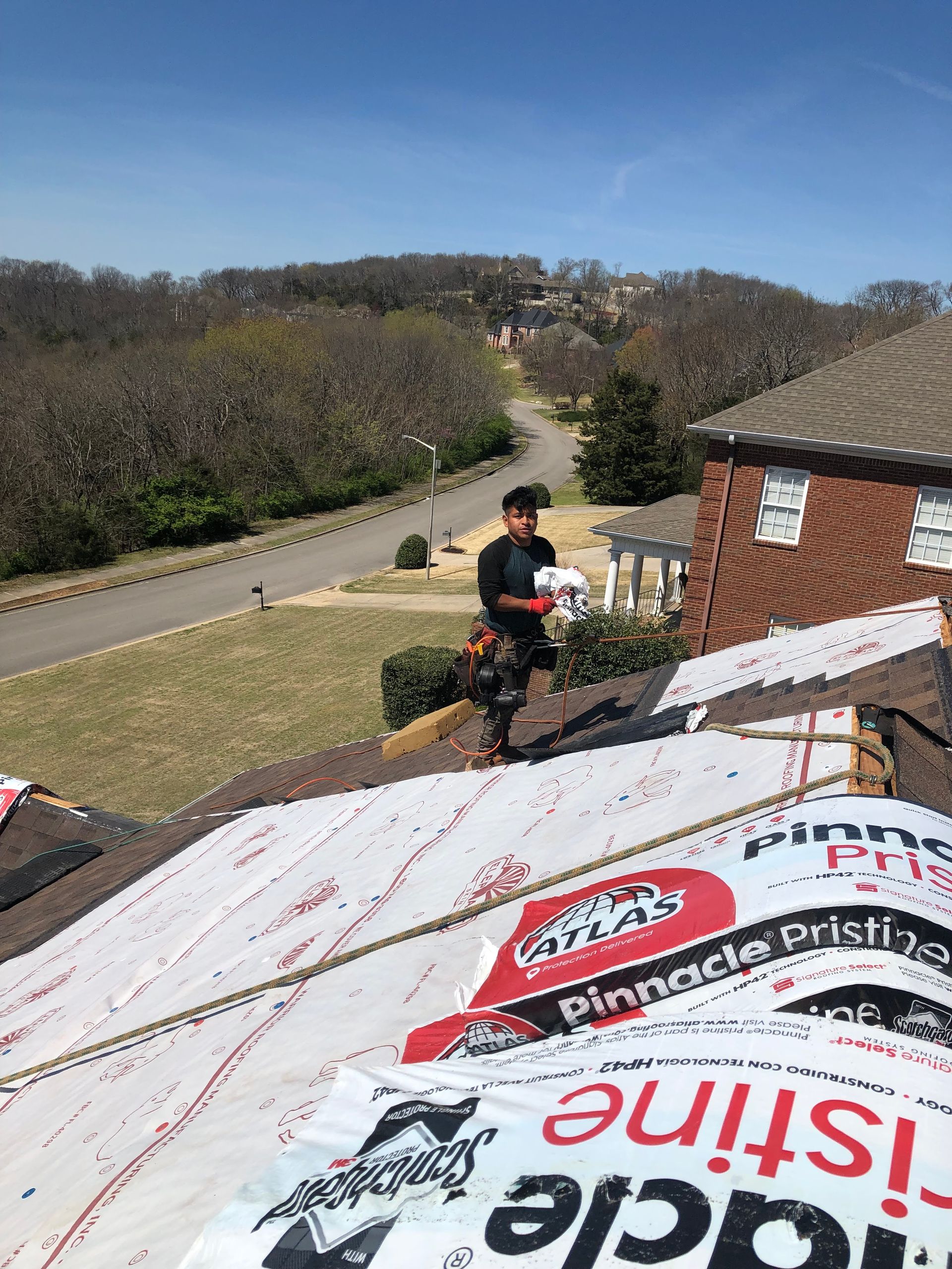 A worker stands on a partially shingled residential roof during a sunny day with a view of a road and trees in the distance.