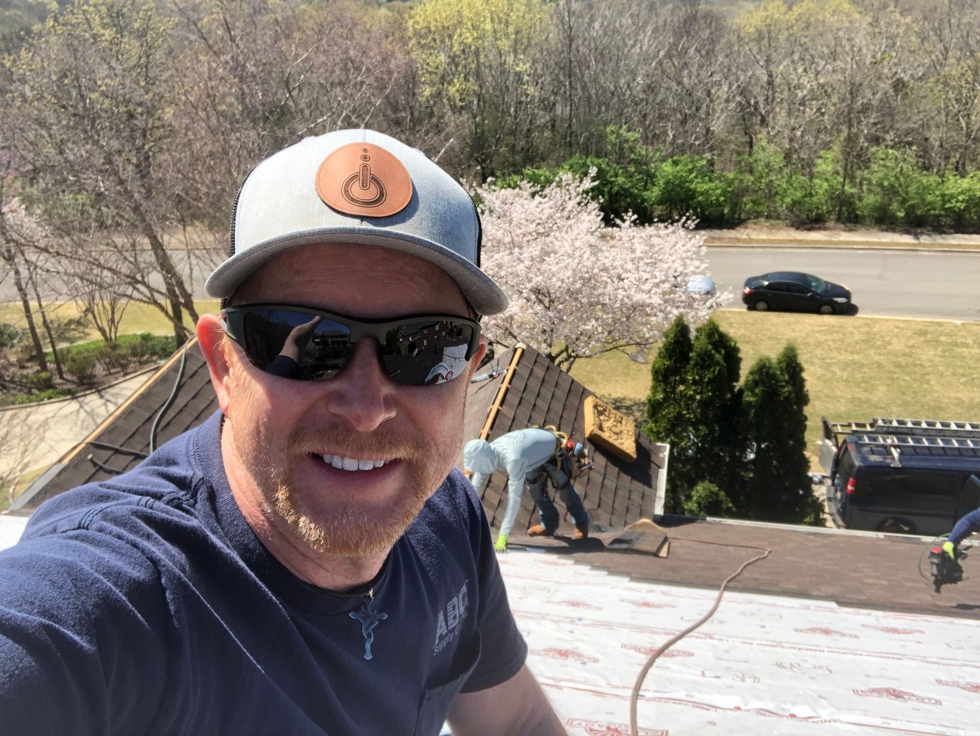 A person wearing sunglasses and a cap smiles for a selfie on a roof under construction, with another worker in the back.