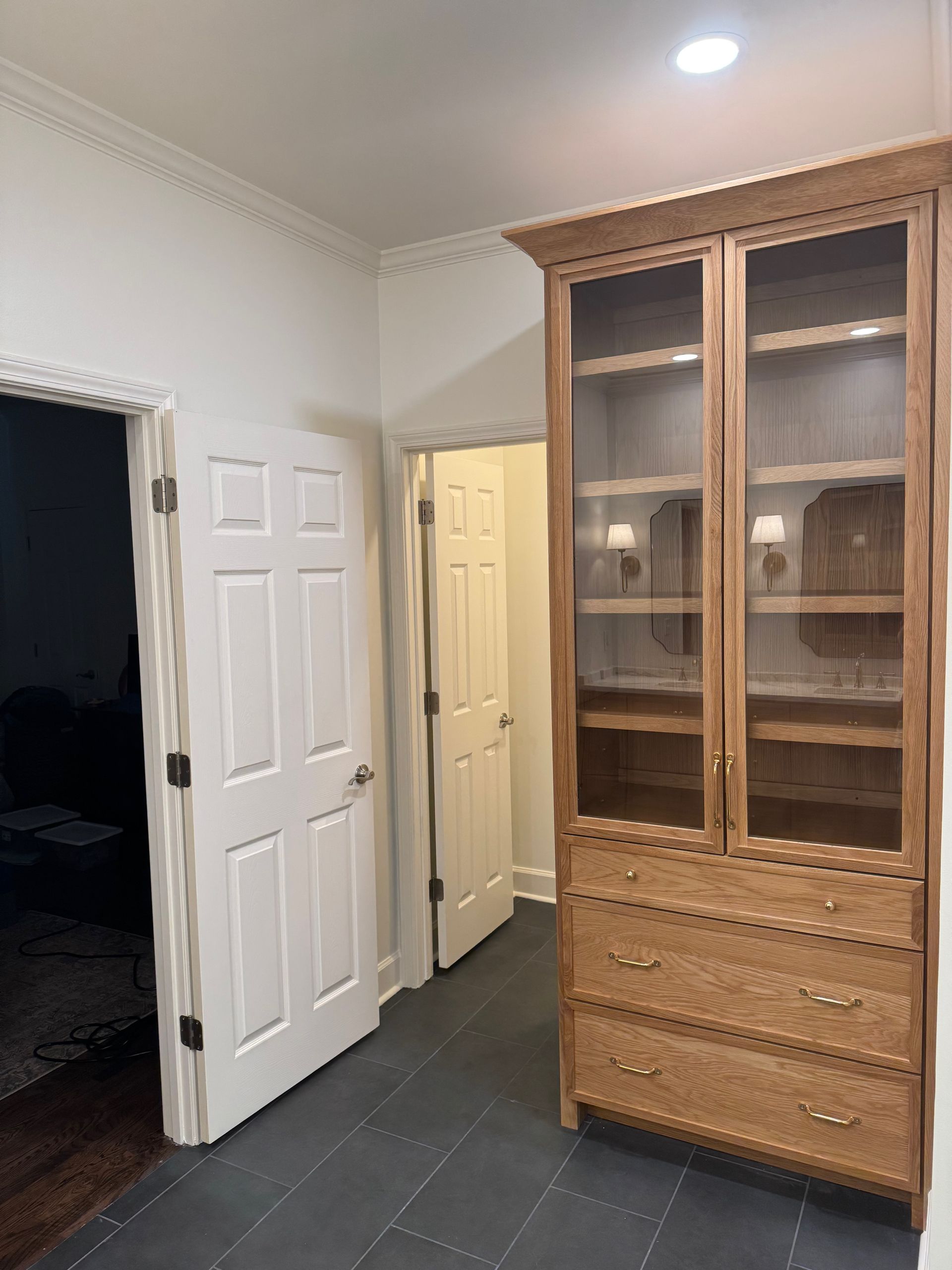 A tall wooden hutch with glass doors and drawers stands on a dark tiled floor next to two white doors in a hallway.