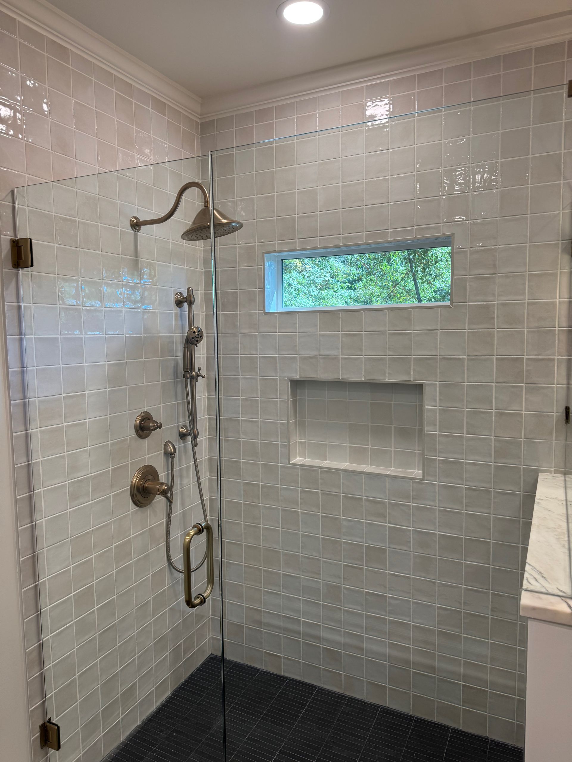 A shower with light gray square tiles, a bronze shower head and handle, a small horizontal window, and a dark tile floor.