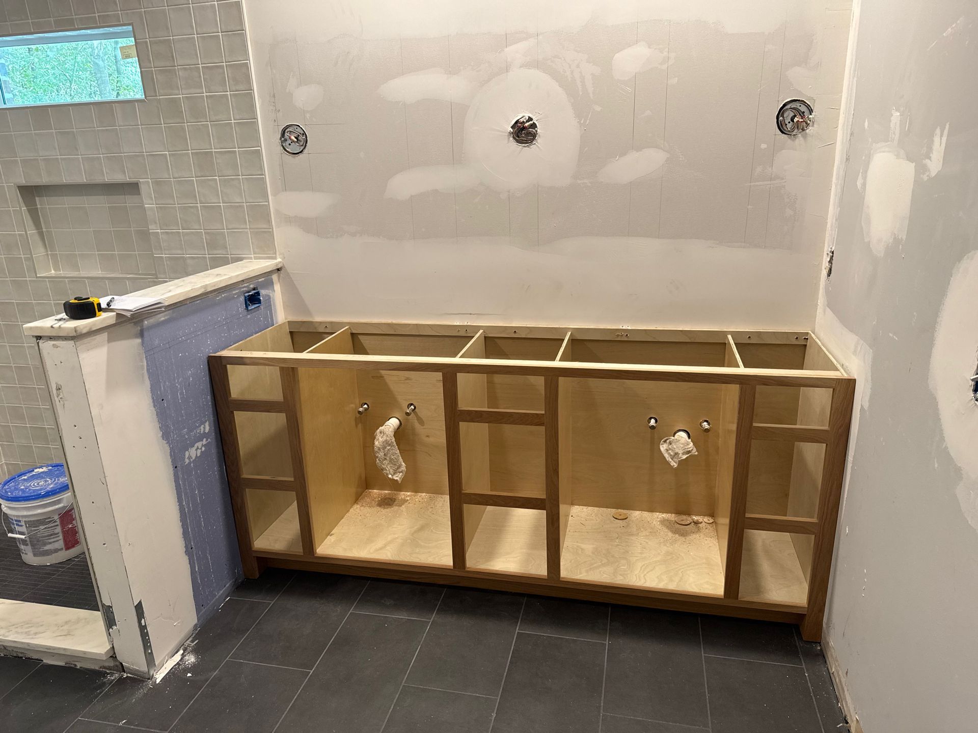 Unfinished bathroom vanity cabinet frame installed against a wall with exposed drywall and plumbing pipes in a room.
