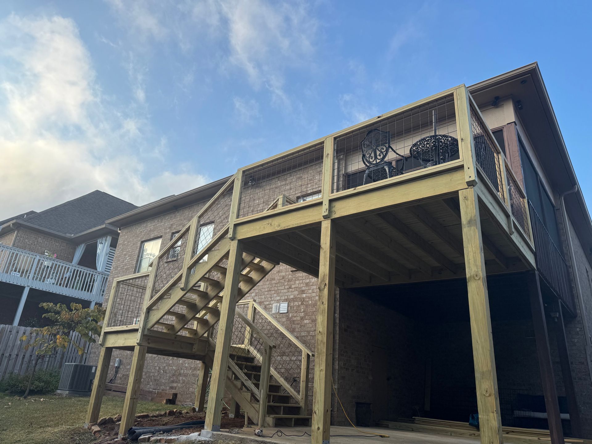 A newly built wooden deck and staircase attached to the side of a brick house under a blue sky.