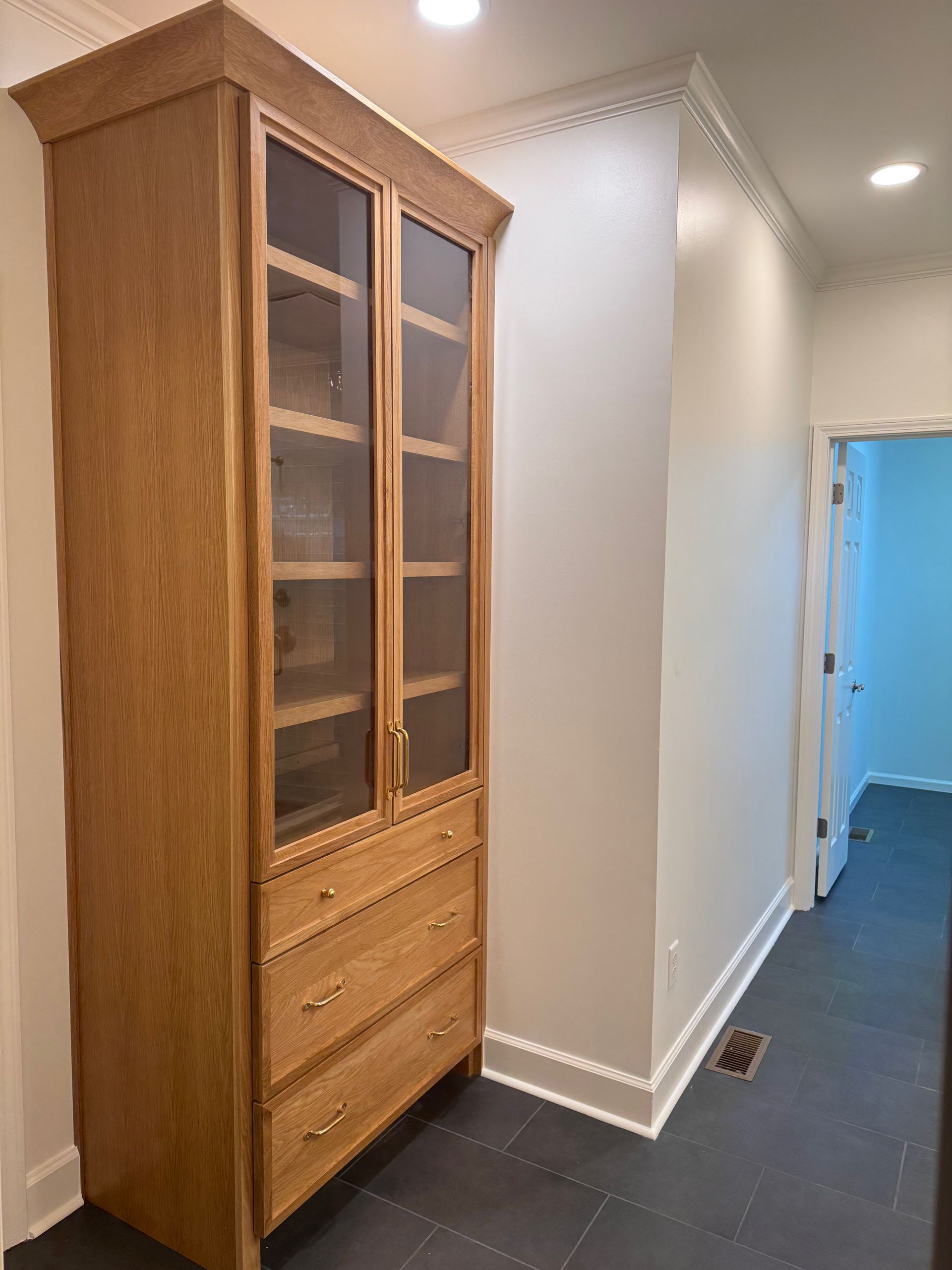 A tall, wood-finished cabinet with glass upper doors and three lower drawers stands on a dark tile floor near a doorway.