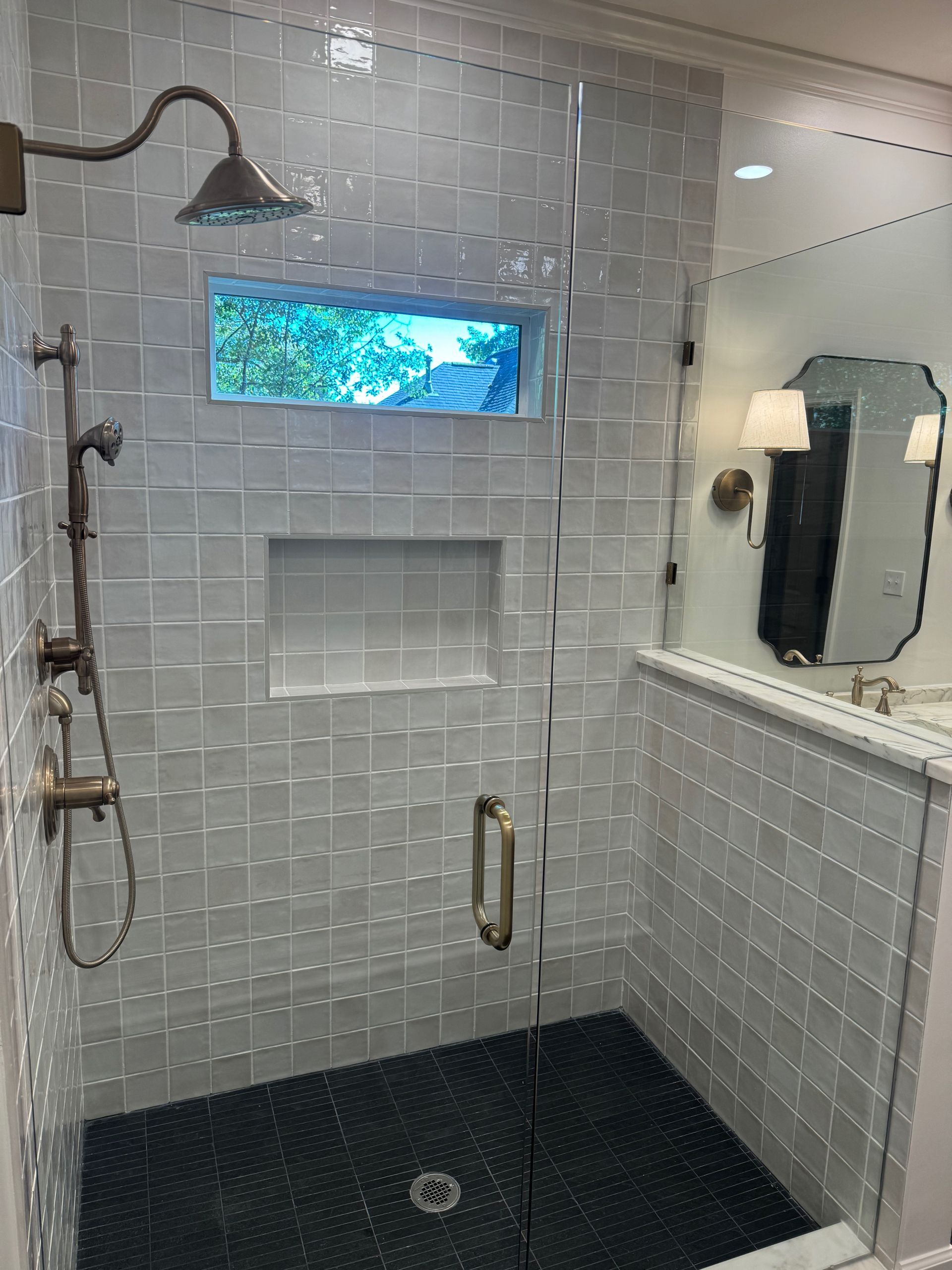 A walk-in shower with white tiled walls, a dark pebble floor, a small window, a bronze showerhead, and a glass door.