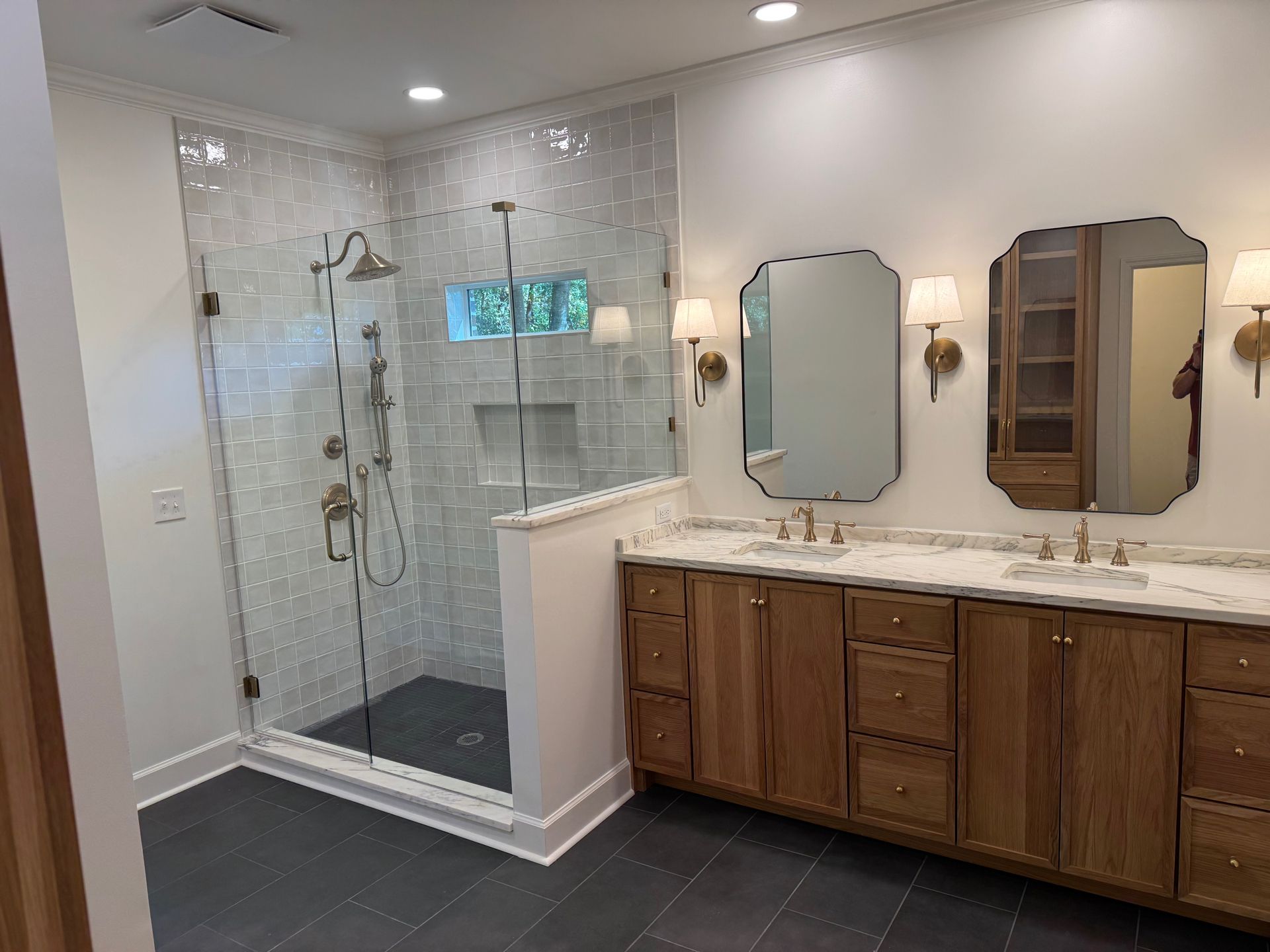 A bathroom with a glass-enclosed shower featuring white subway tiles, a wooden vanity, and dual mirrors with wall sconces.