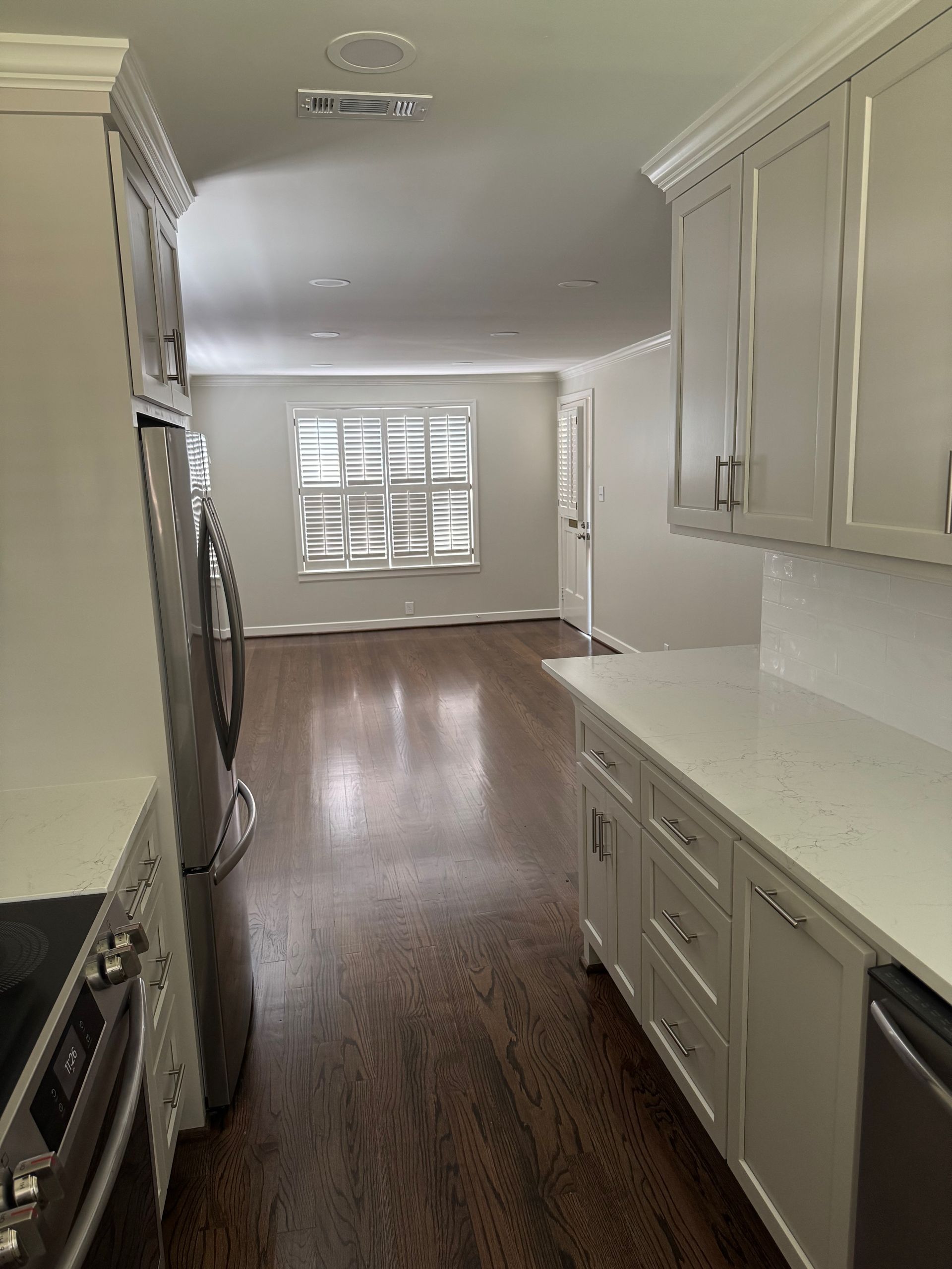 A bright kitchen with light grey cabinets, stainless steel appliances, and dark wood floors, looking into an open room.