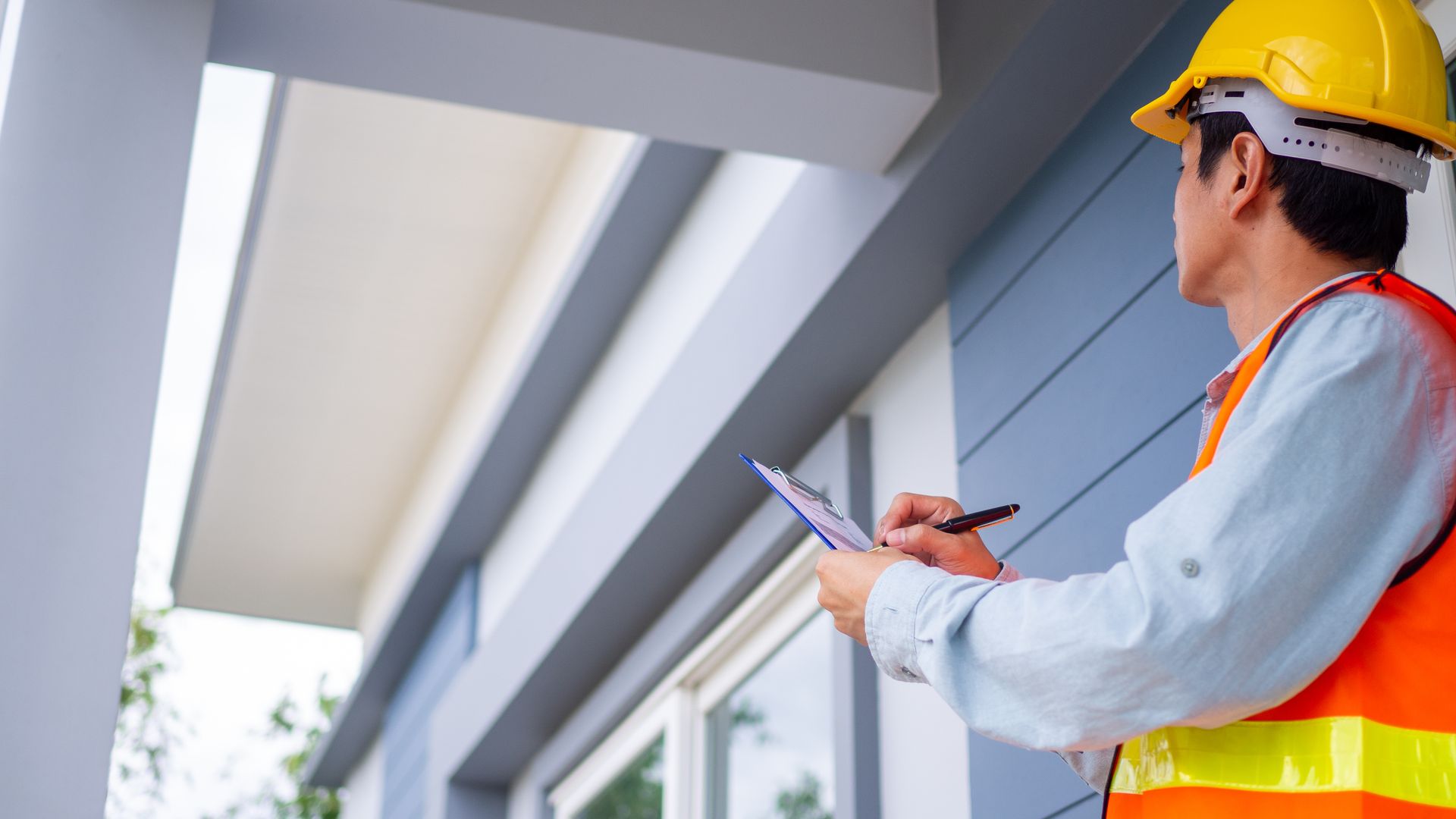 A professional wearing a yellow hard hat and orange safety vest inspects a building exterior while writing on a clipboard.