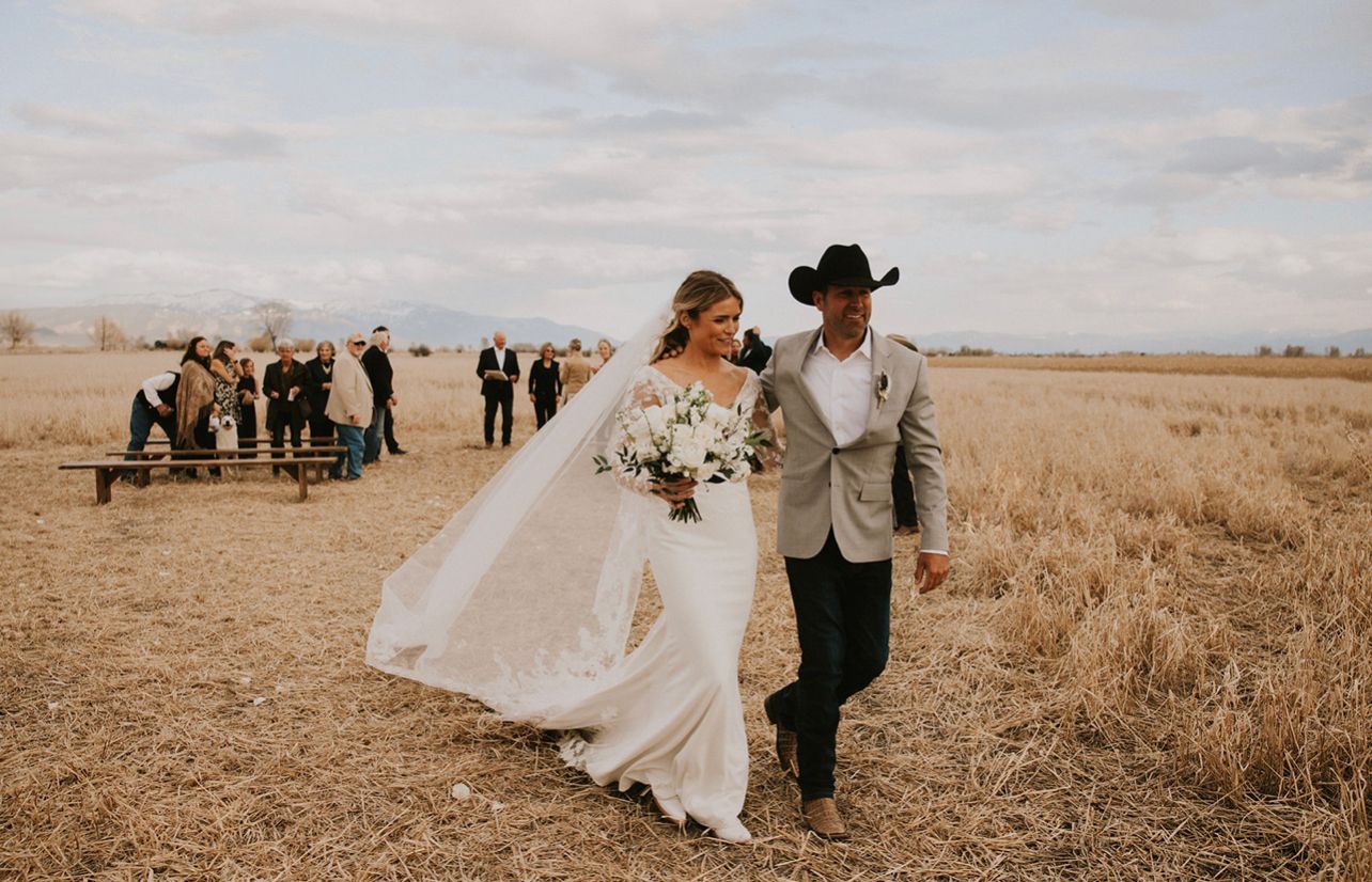 A bride and groom are walking through a field holding hands.