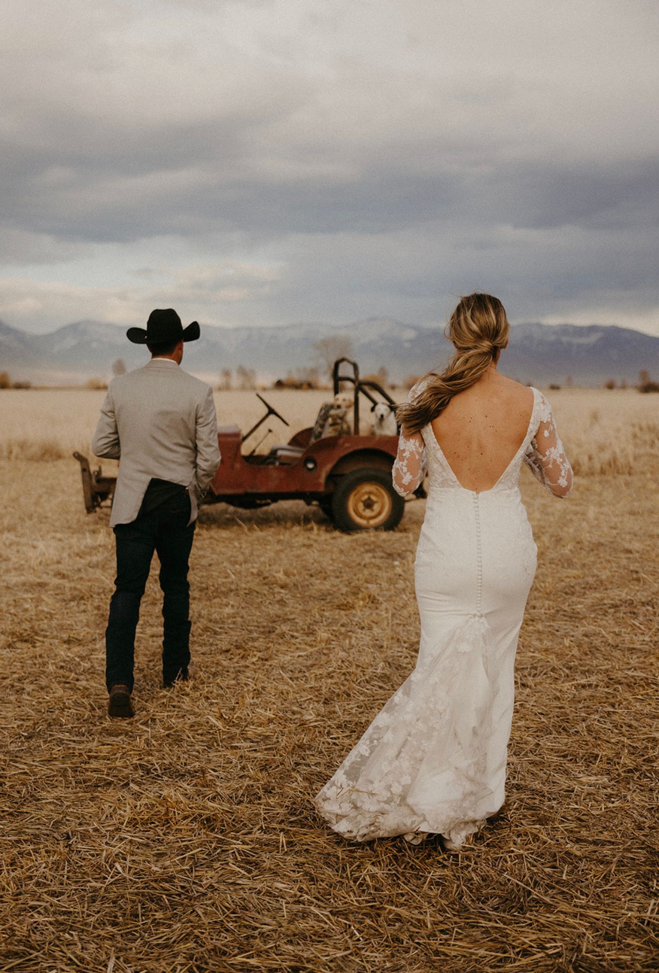 A bride and groom are walking in a field with a jeep in the background.