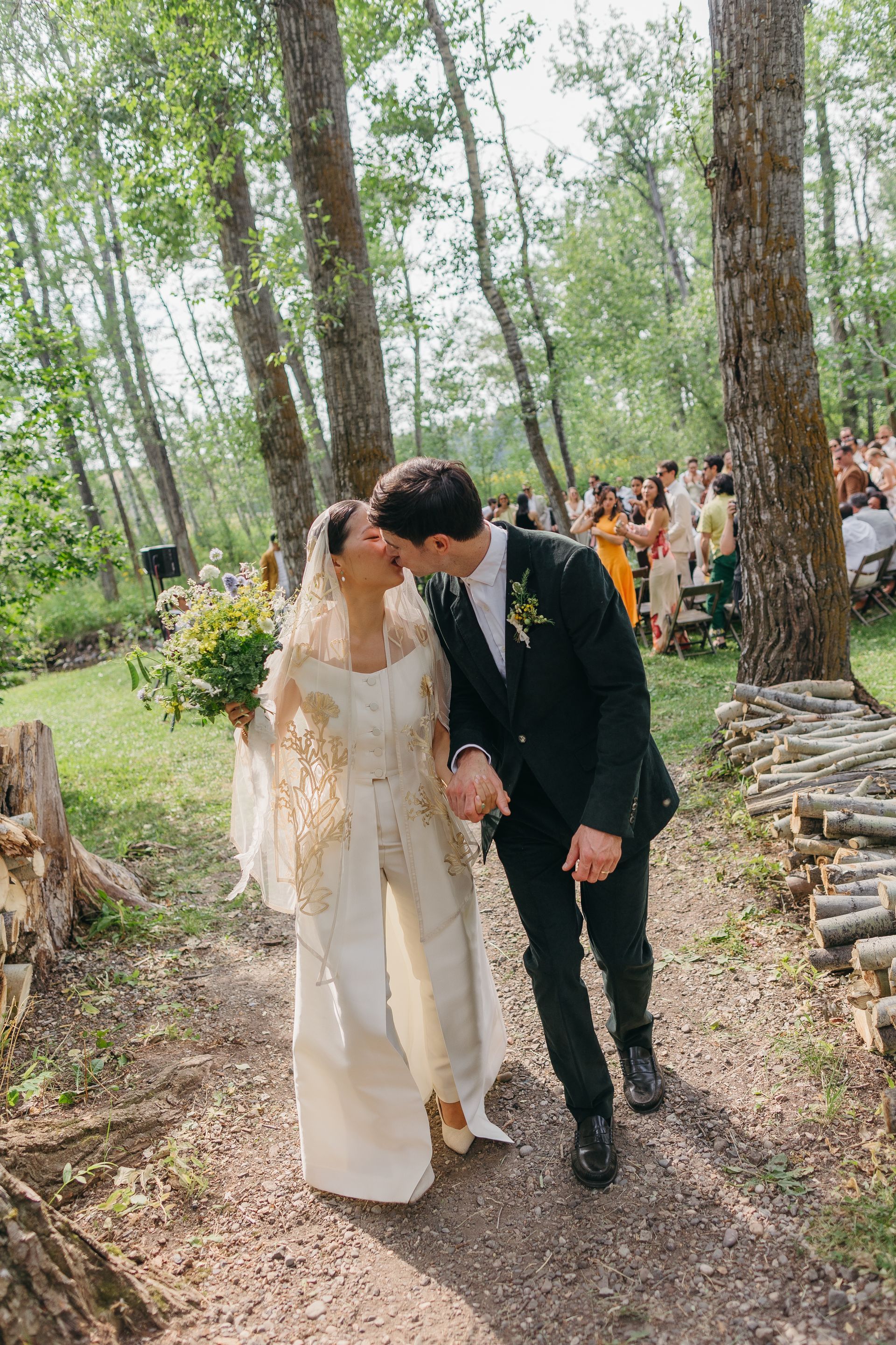 A bride and groom are kissing while walking down a path in the woods.