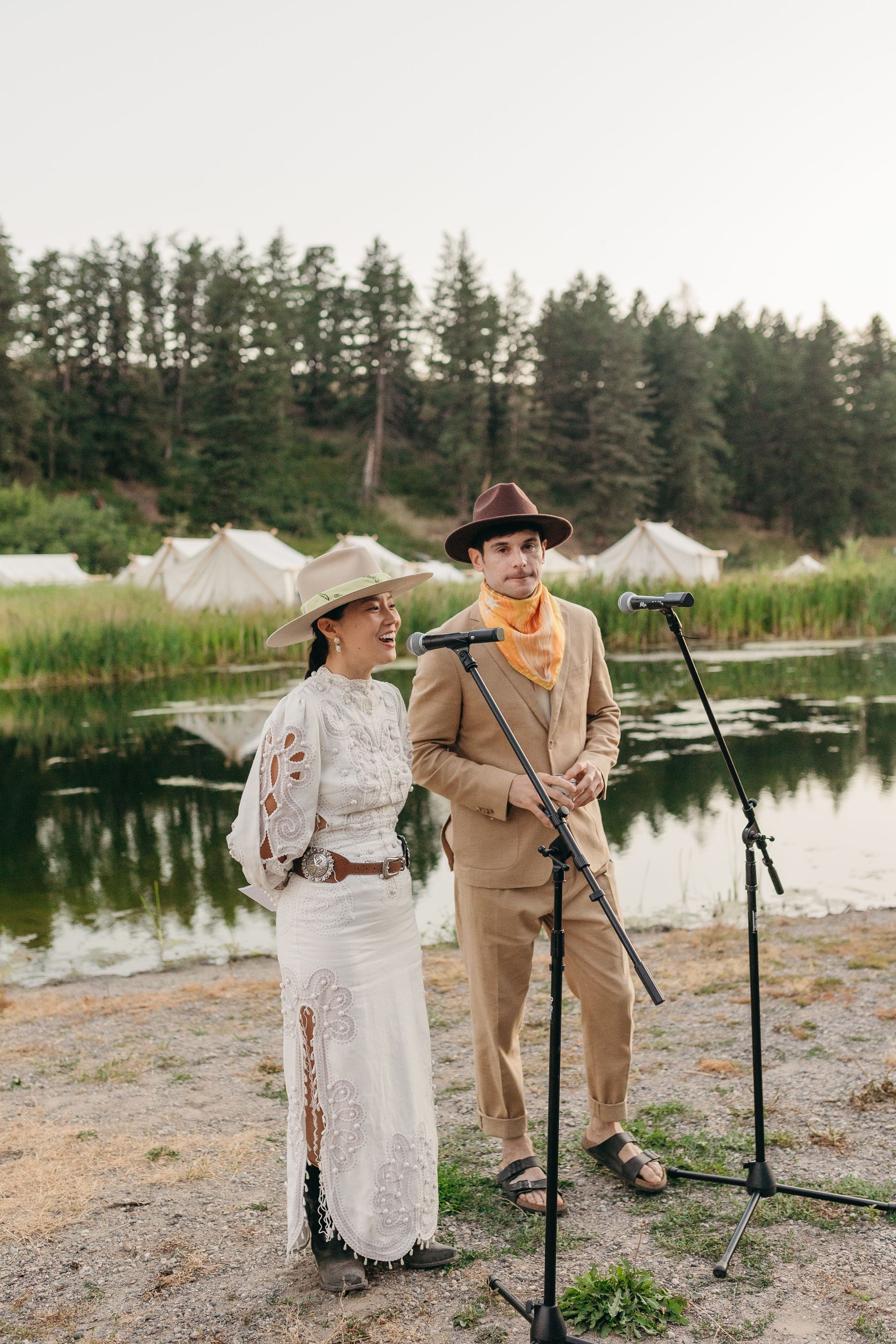 A bride and groom are singing into microphones in front of a lake.