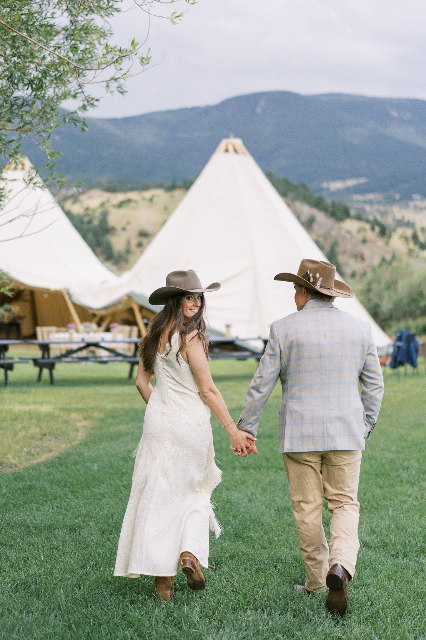 A bride and groom are walking in a field holding hands in front of a tent.