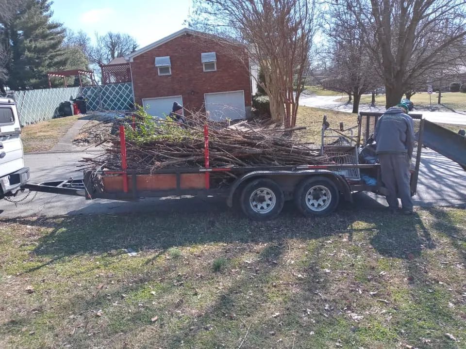 A man is pushing a trailer full of logs in front of a brick house.