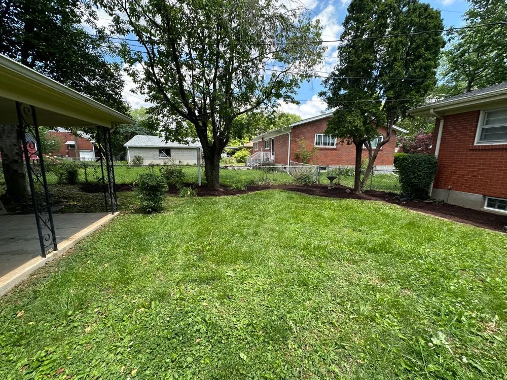 A lush green backyard with a covered porch and a brick house in the background.