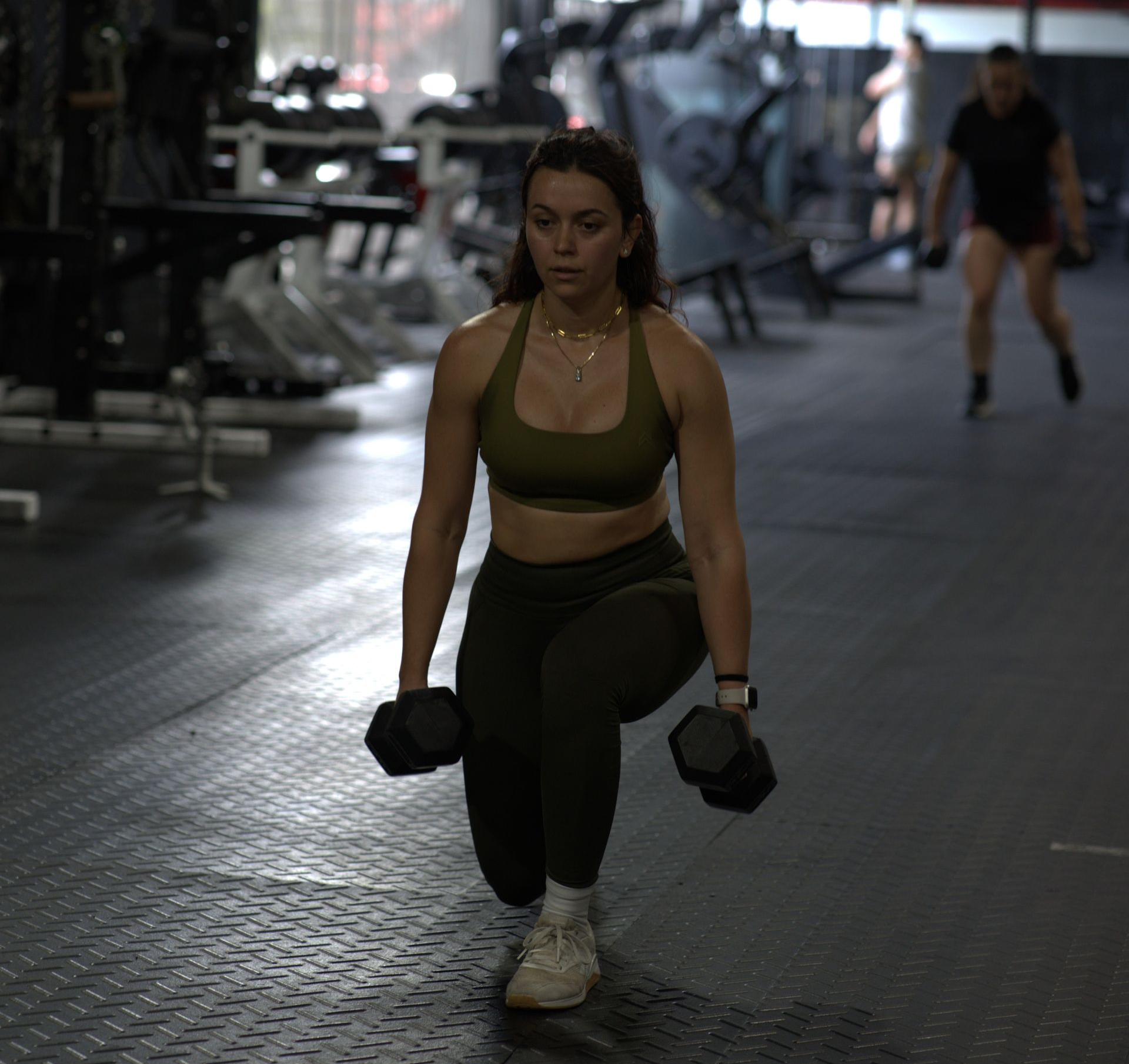 Woman doing dumbbell lunges at The LOX gym in Doral, FL. She's in olive-colored workout attire, with focused expression.