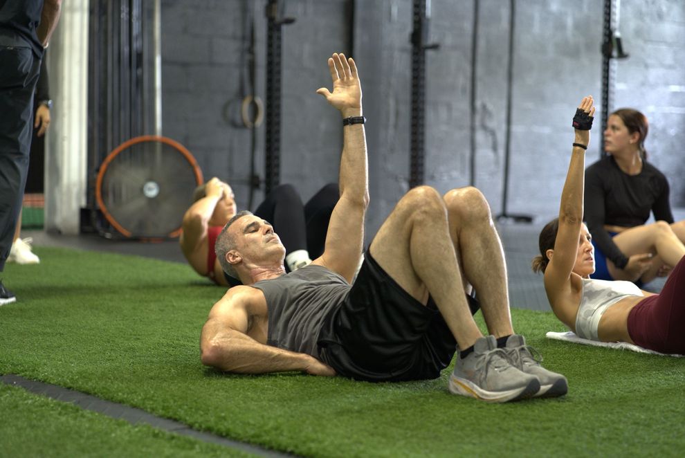 People doing sit-ups on green turf at The LOX gym in Doral, FL. Man raises hand. Others in the background.