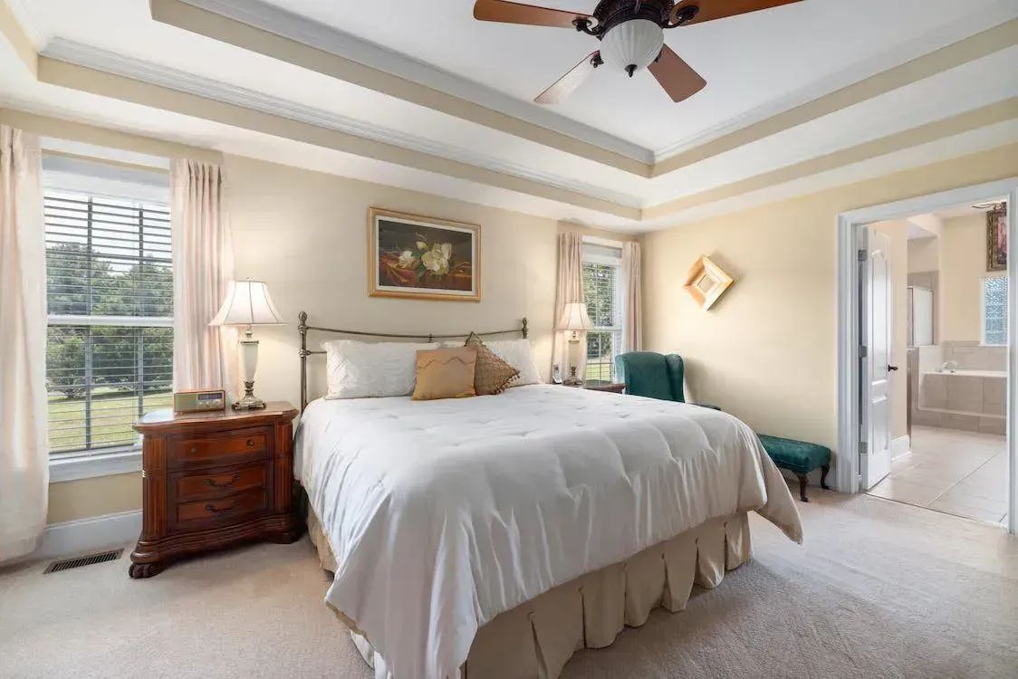 Bedroom with bed, nightstands, windows, and access to a bathroom. Beige walls, carpet, and ceiling beams.