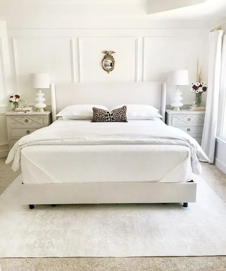 White bedroom with bed, bedside tables, rug, decorative wall panels, and an ornate mirror.