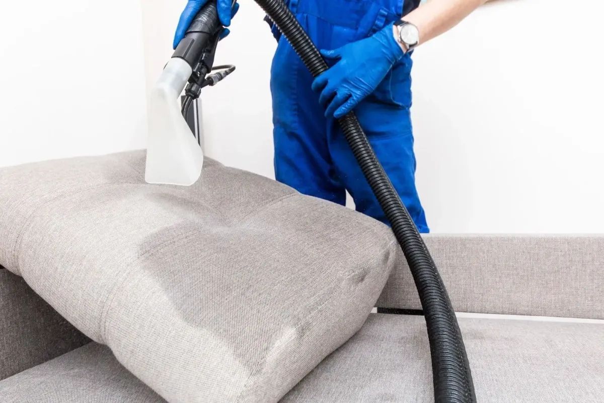 Person in blue overalls and gloves cleaning a gray sofa cushion with a vacuum.