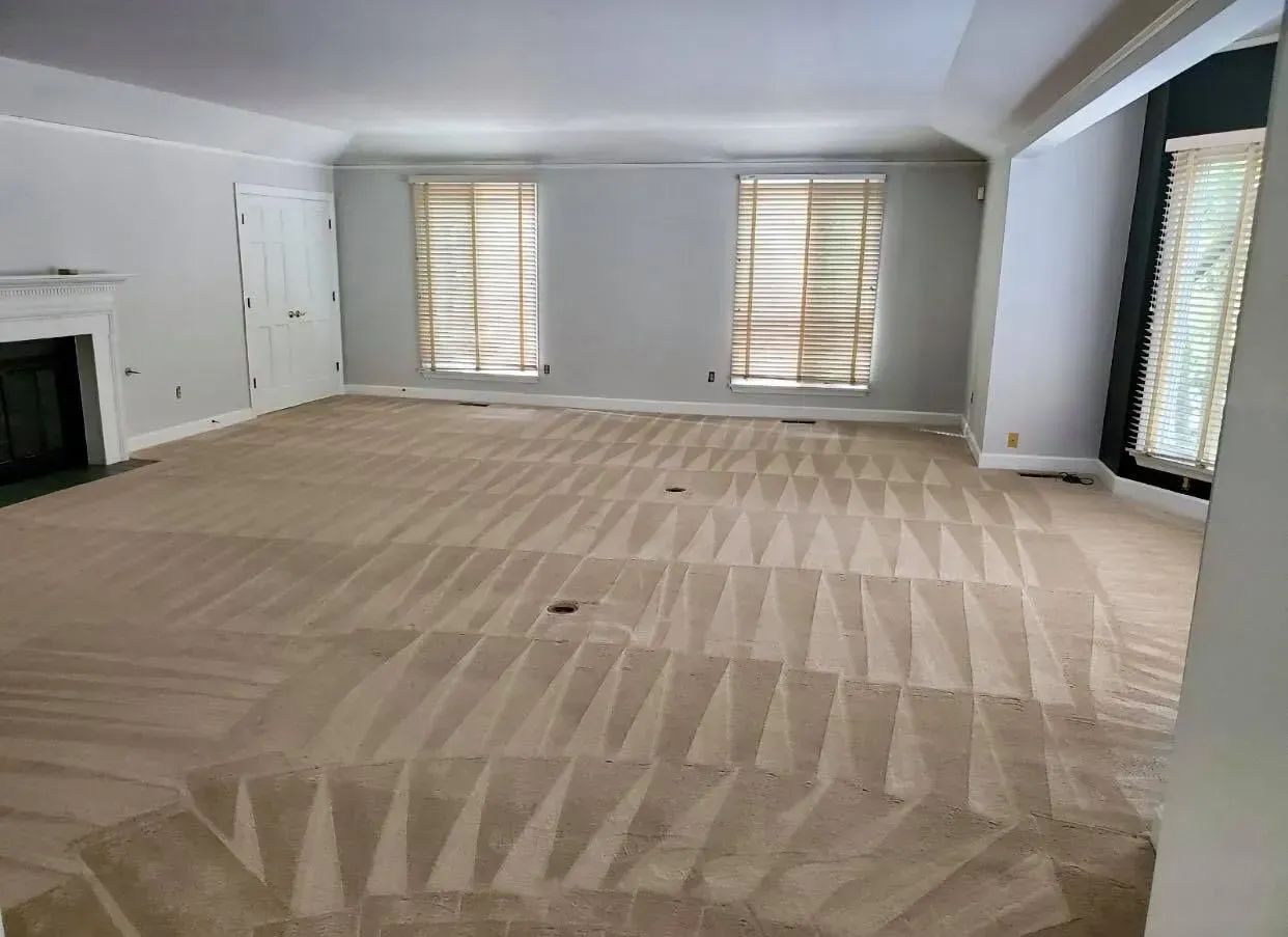 An empty room with light gray walls and beige carpet in a geometric pattern. Two windows with shades.