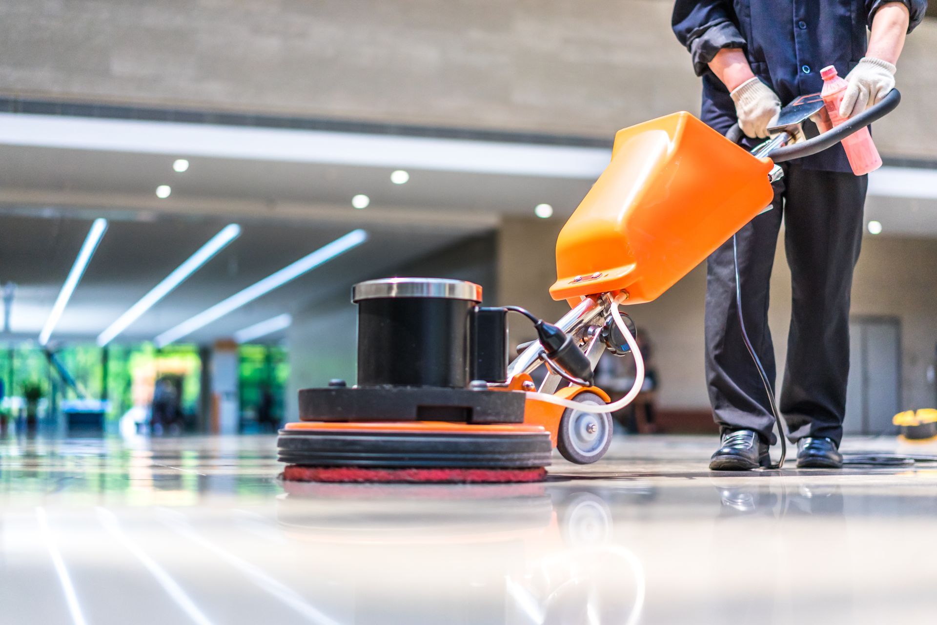 Person using an orange and black floor buffer machine on a polished floor in a building.
