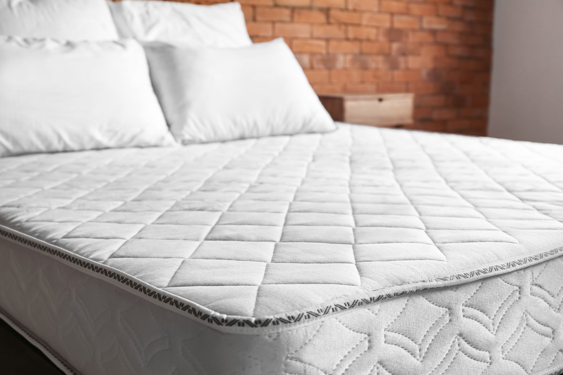 White mattress with stitched pattern and pillows on a bed in front of a brick wall.