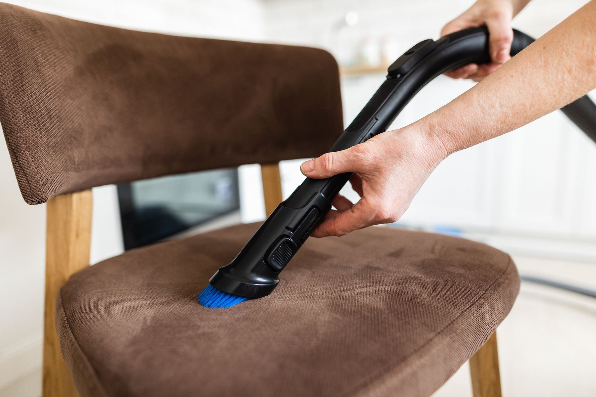 Person vacuuming a brown upholstered chair with a blue brush attachment.