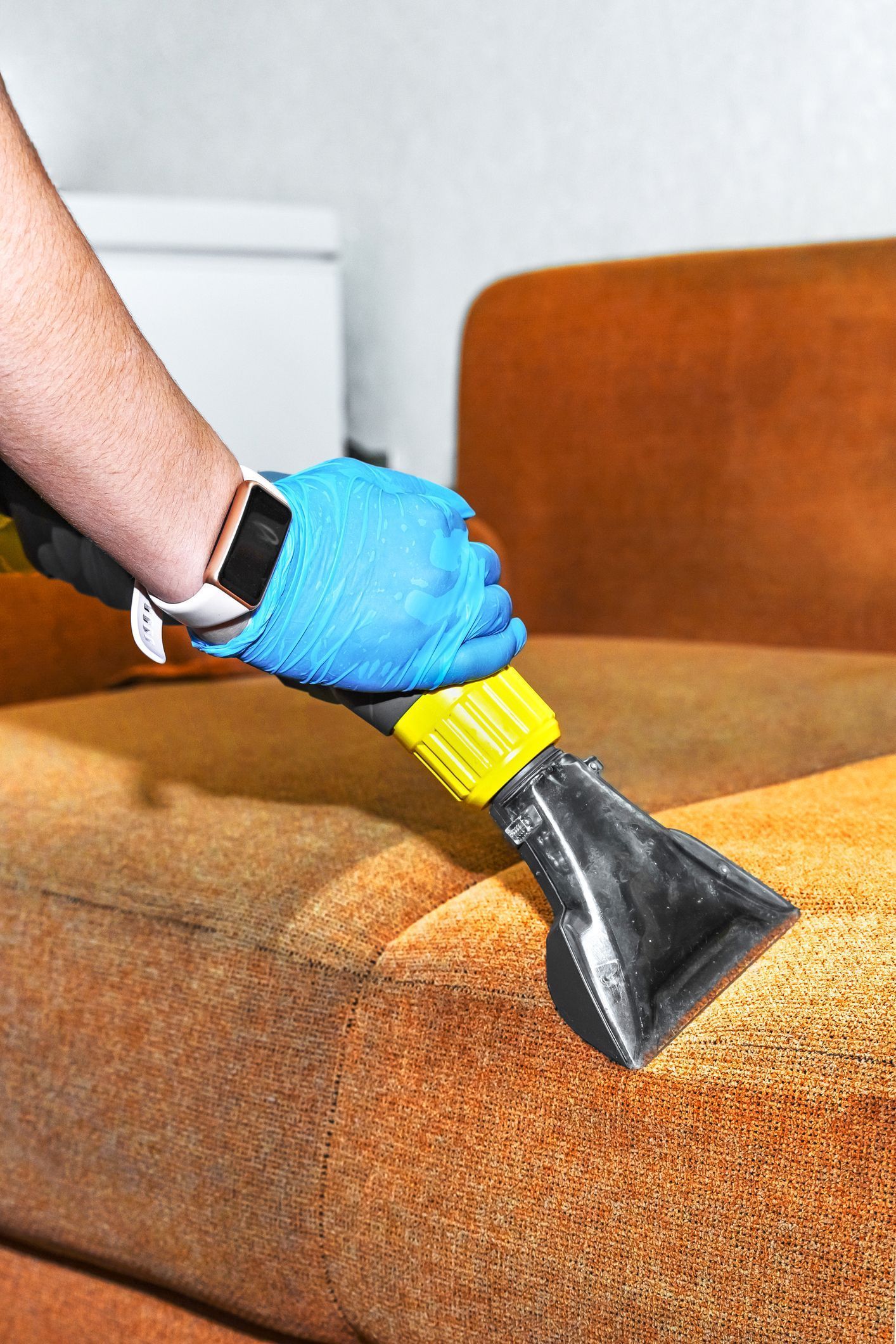 Person wearing blue gloves cleans an orange couch with a cleaning machine.