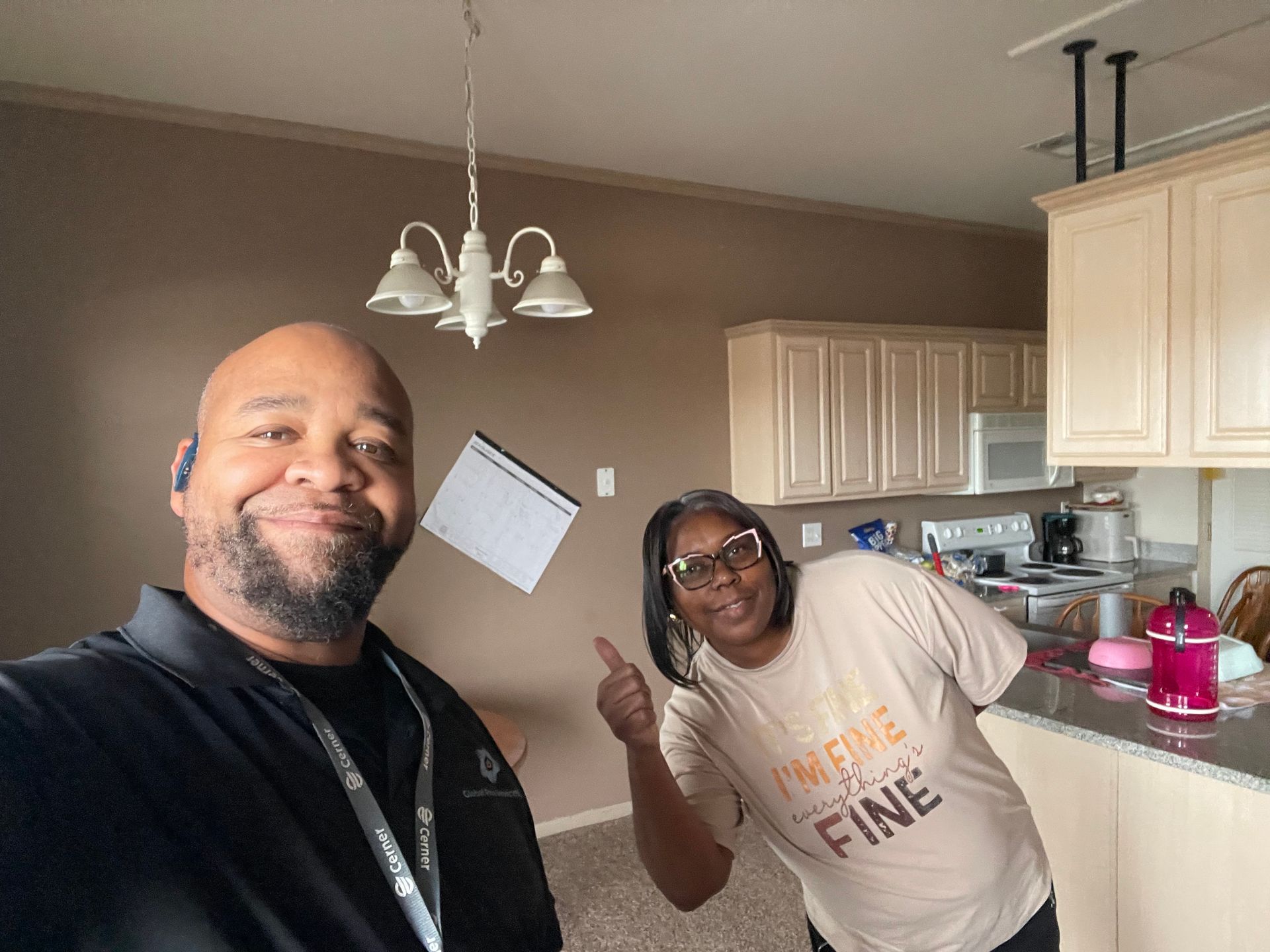 Two people pose for a selfie in an empty kitchen with beige walls and cabinets, with one person giving a thumbs up.