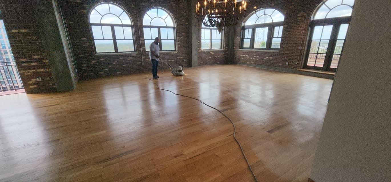 A person refinishing a wood floor in a large room with arched windows and a brick wall.