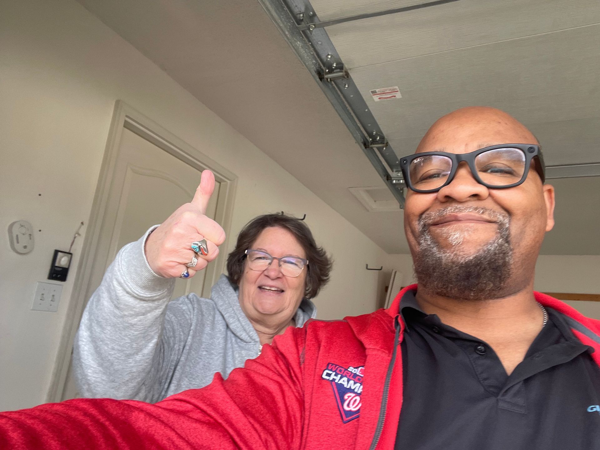 Two people posing for a selfie in a garage; one wears a red jacket and glasses, the other gives a thumbs up.