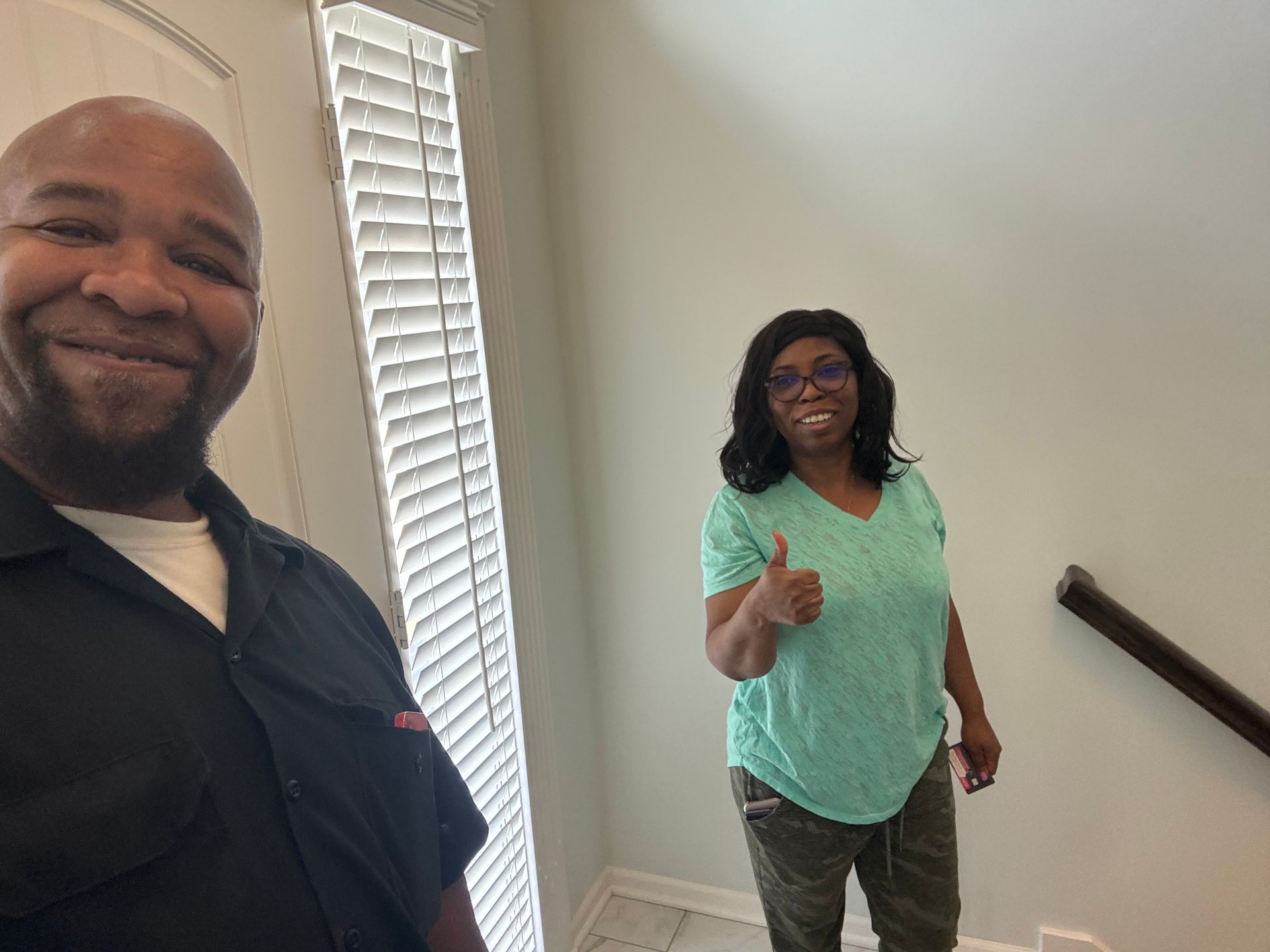 A person smiling for a selfie with another person in the background giving a thumbs-up by a staircase and window.