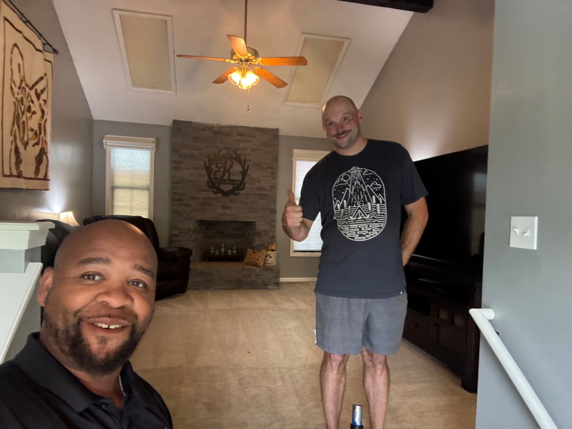 Two men in a living room, one smiling in the foreground and one giving a thumbs-up by a fireplace in the background.
