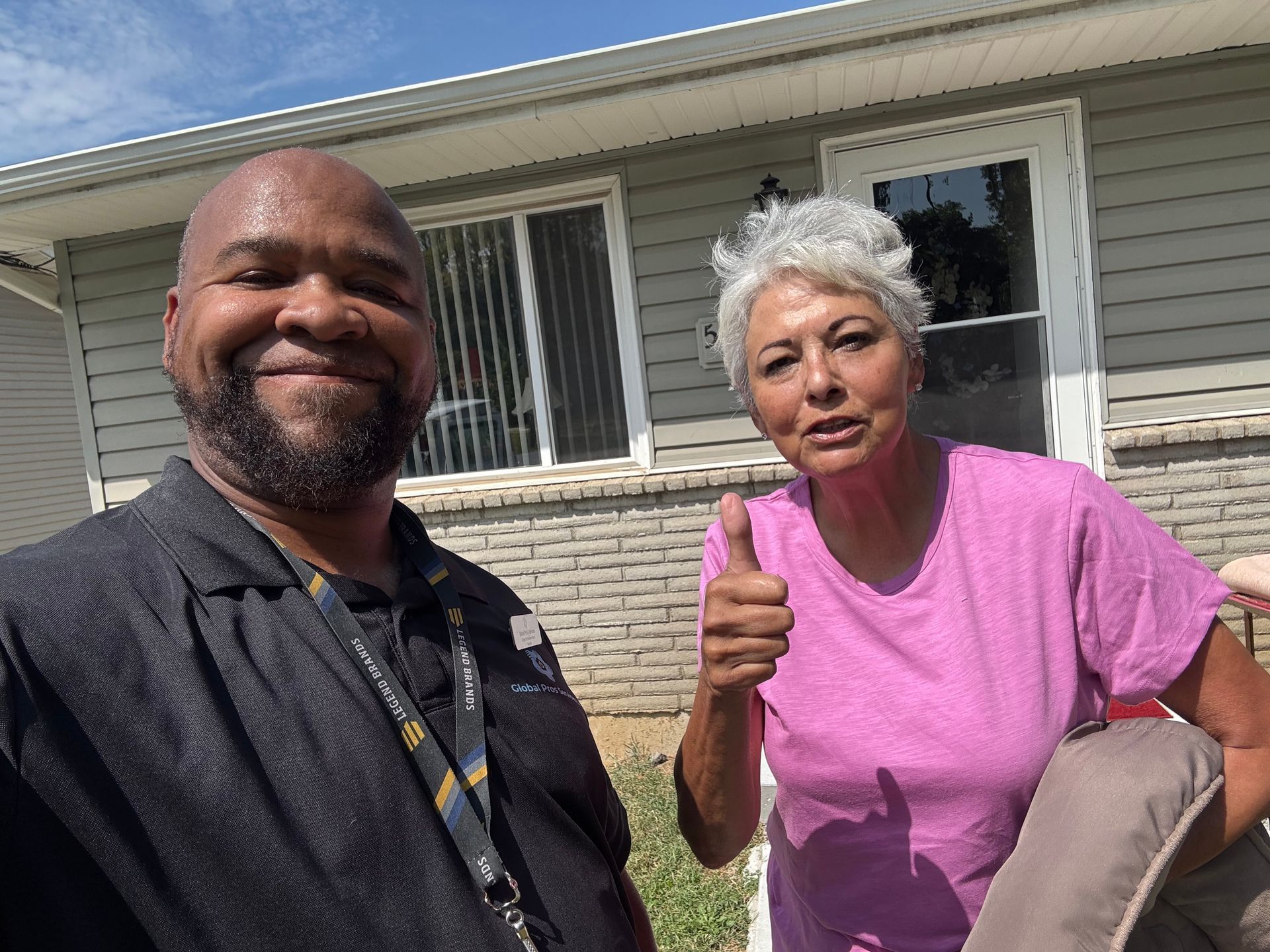 A person in a black polo shirt smiles next to a person in a pink shirt giving a thumbs up in front of a house.