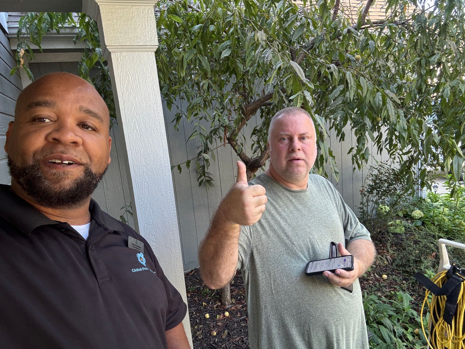 A man in a black polo shirt stands with another man who is giving a thumbs-up while holding a small black device outside.