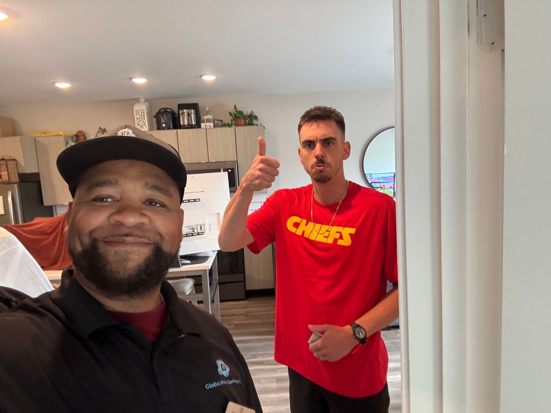 A man in a black polo smiles in the foreground, while a man in a red Chiefs t-shirt gives a thumbs-up in the background.