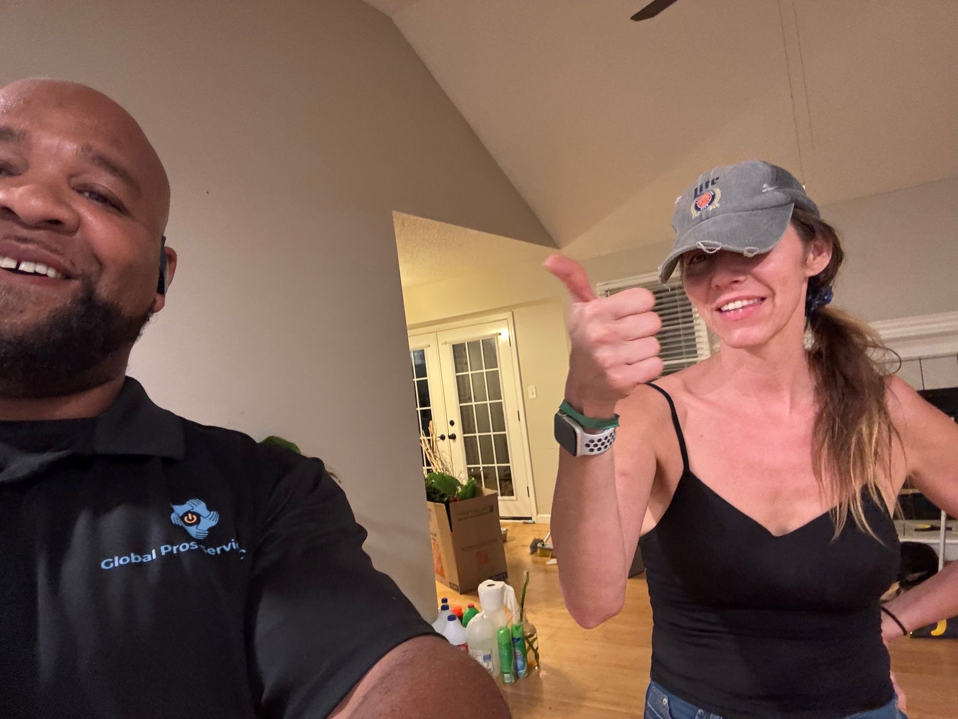 Two people standing indoors, one smiling and the other giving a thumbs-up gesture while wearing a baseball cap.