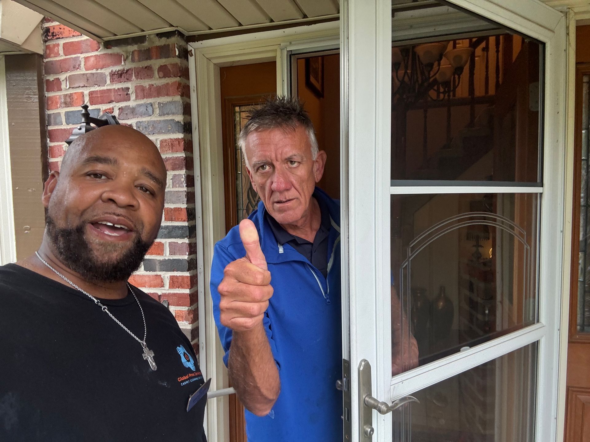 Two individuals stand at a brick house entrance, one smiling in a black shirt, the other in a blue jacket giving a thumbs up.