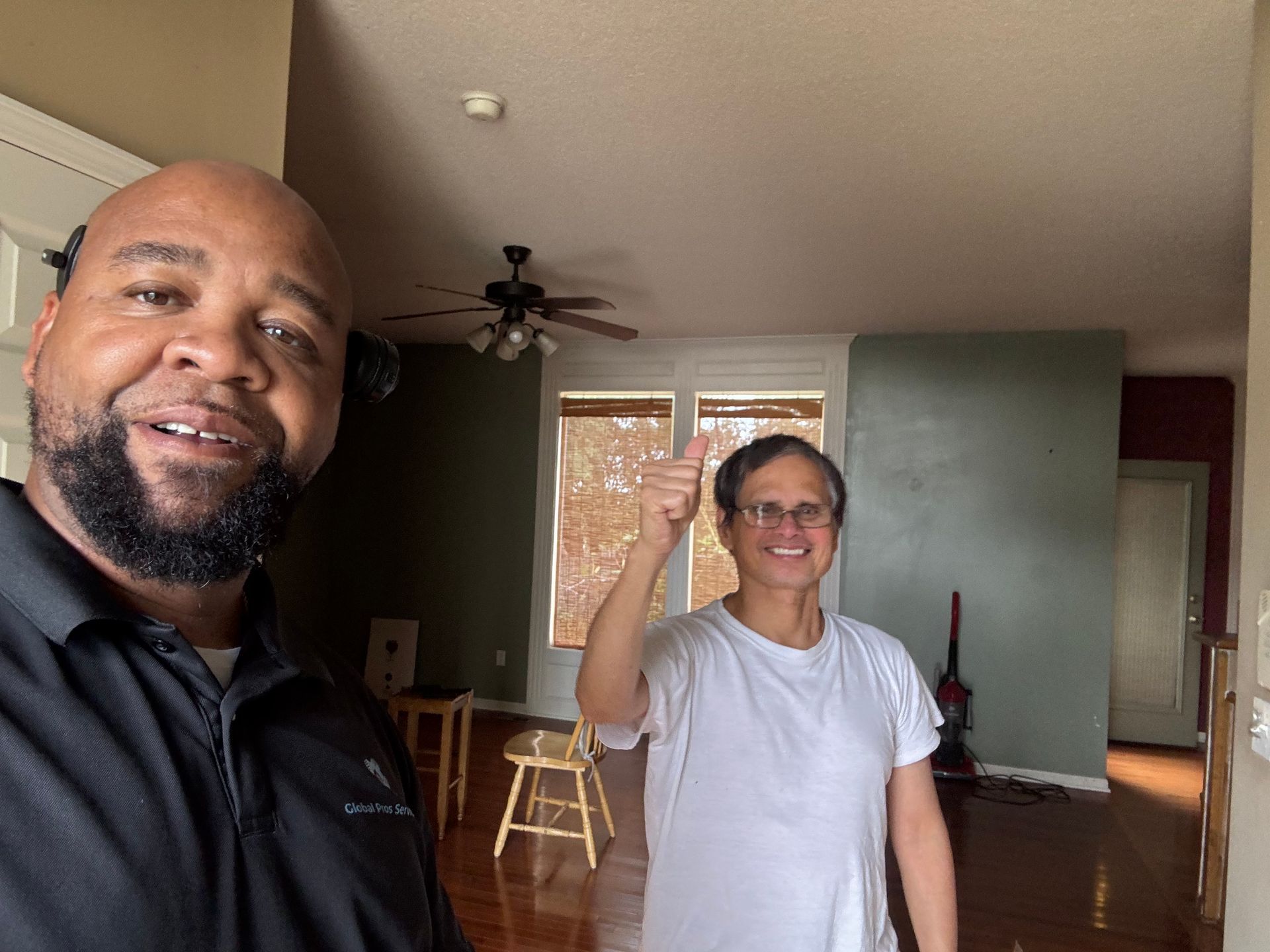A person in a black polo smiles beside someone in a white t-shirt who is giving a thumbs up in an empty apartment room.