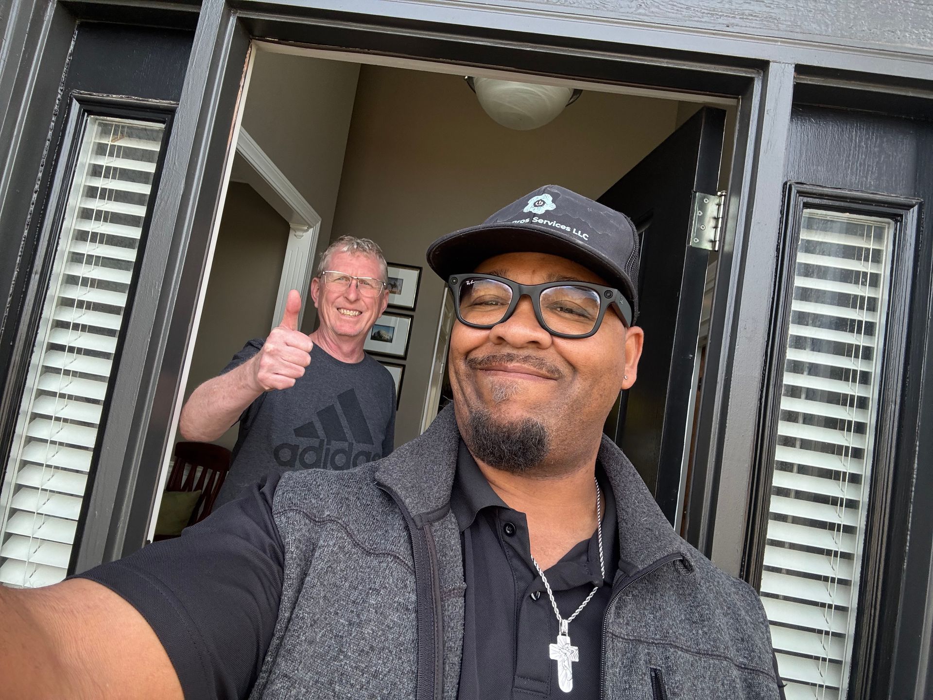 A person wearing a hat and glasses takes a selfie in a doorway while another person in the background gives a thumbs up.