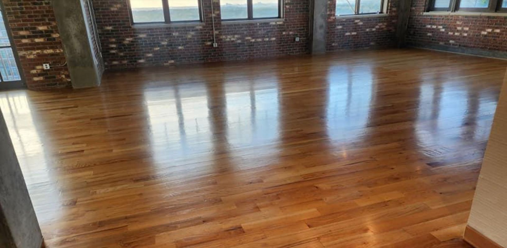 Reflective hardwood floor in a room with exposed brick walls and large windows.
