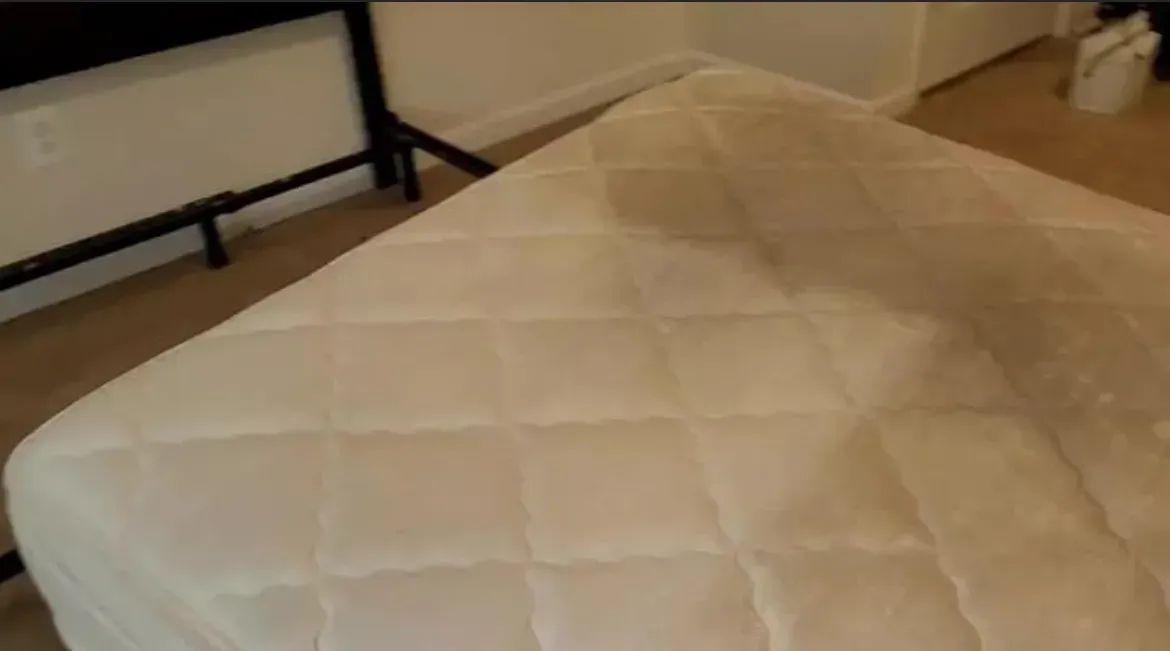 White quilted mattress on a bed frame, in a room with light flooring and a partially visible wall.