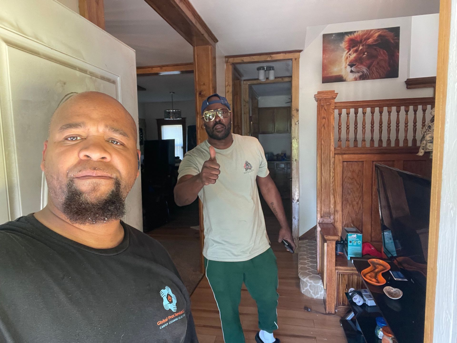 Two men in a living space; one in the foreground takes a selfie while the other stands behind, giving a thumbs up.