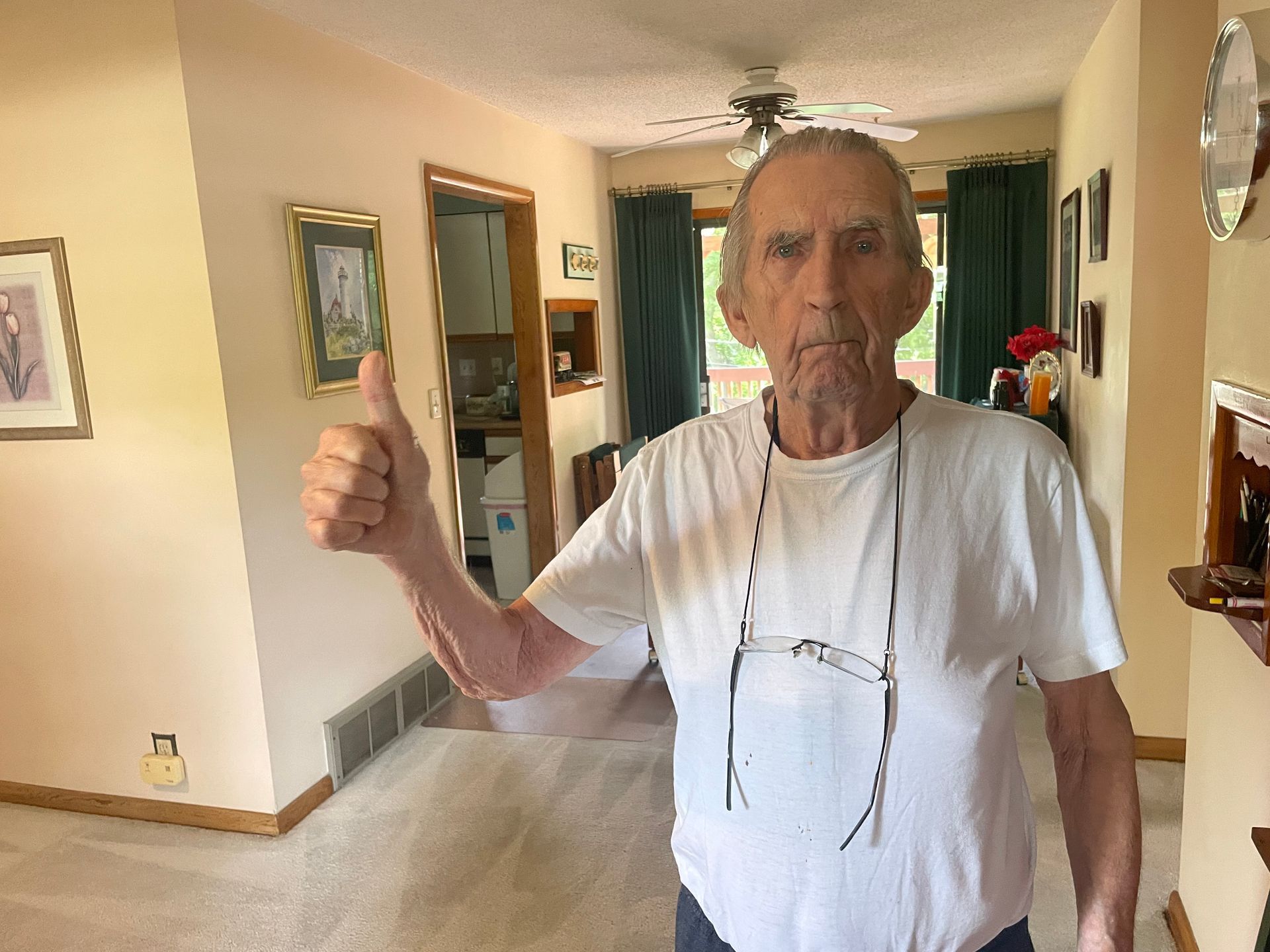 A person in a white t-shirt stands in a living room, smiling and giving a thumbs up gesture.