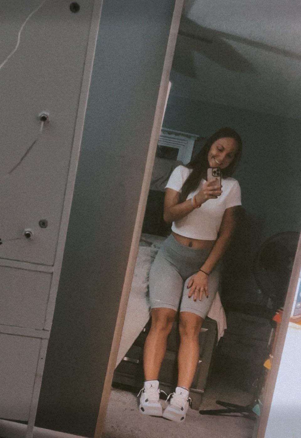 A person wearing a white t-shirt and grey biker shorts stands in front of a mirror, smiling while taking a selfie.