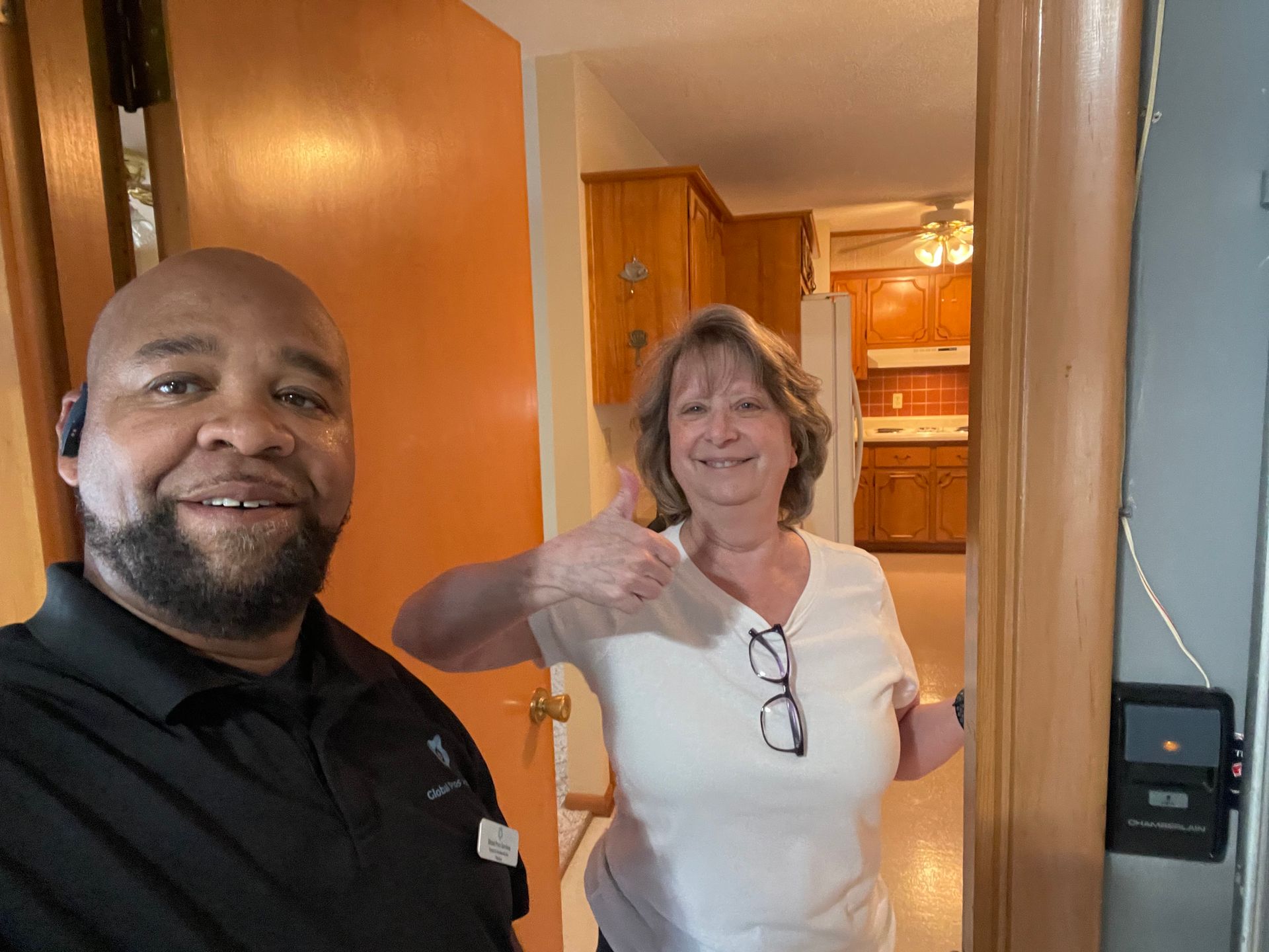 Two people posing for a photo in a doorway; one gives a thumbs-up, with a kitchen visible in the background.
