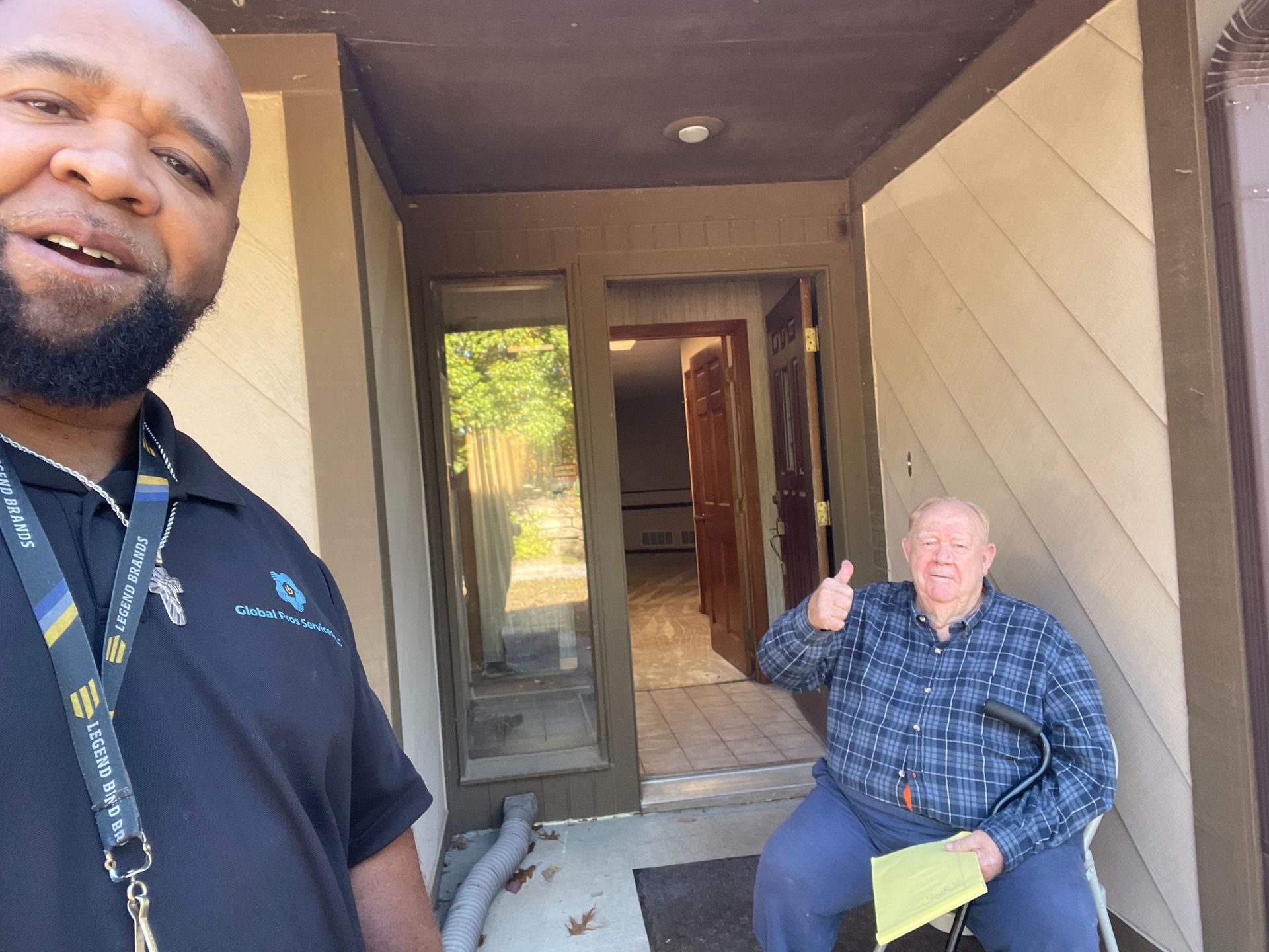 A smiling man stands in the foreground, while a man seated with a cane gives a thumbs up outside an open doorway.