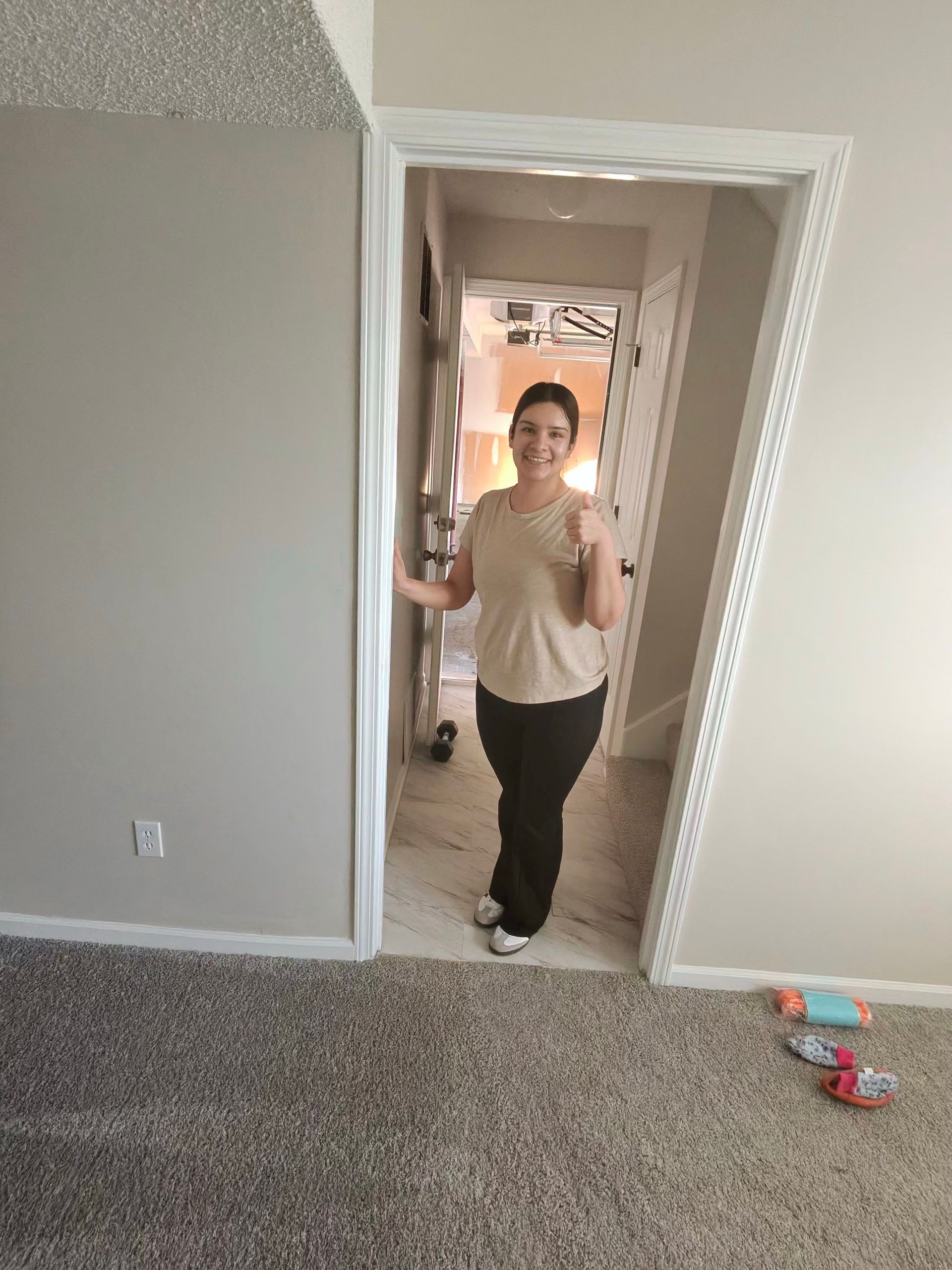 A person stands in a doorway wearing a beige top and black pants, smiling and giving a thumbs-up gesture.