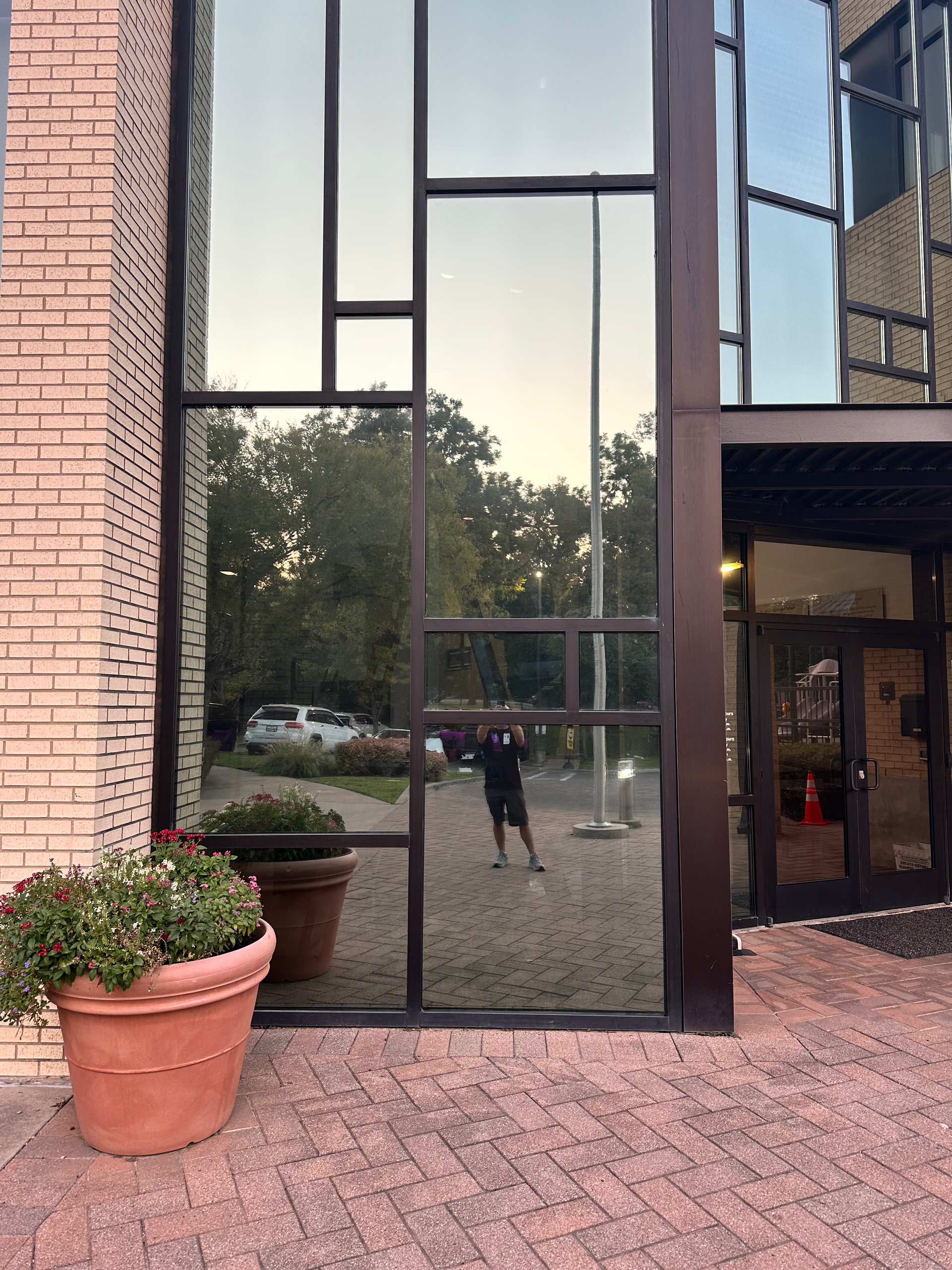 A person reflected in a large window with trees, cars, and a brick building visible. A flower pot is on the left.