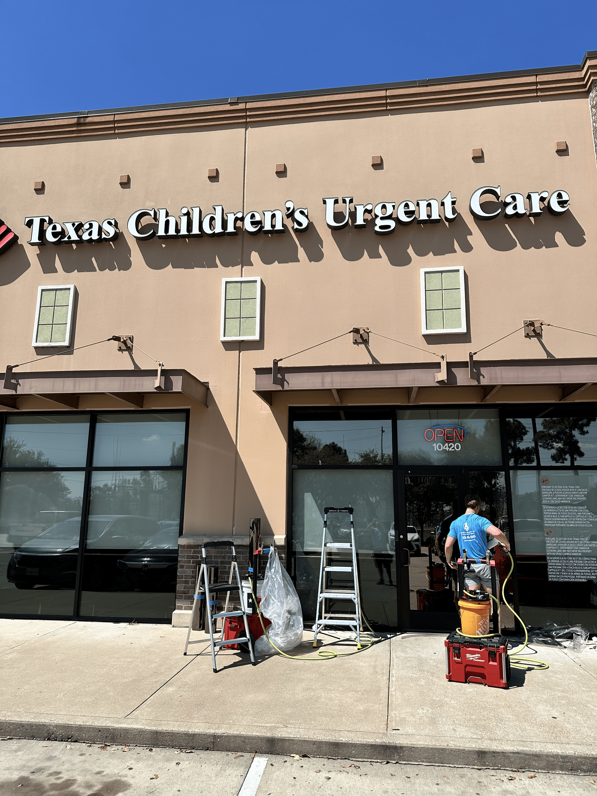 Texas Children's Urgent Care exterior; construction worker, ladder, and tools. Beige building, blue sky.